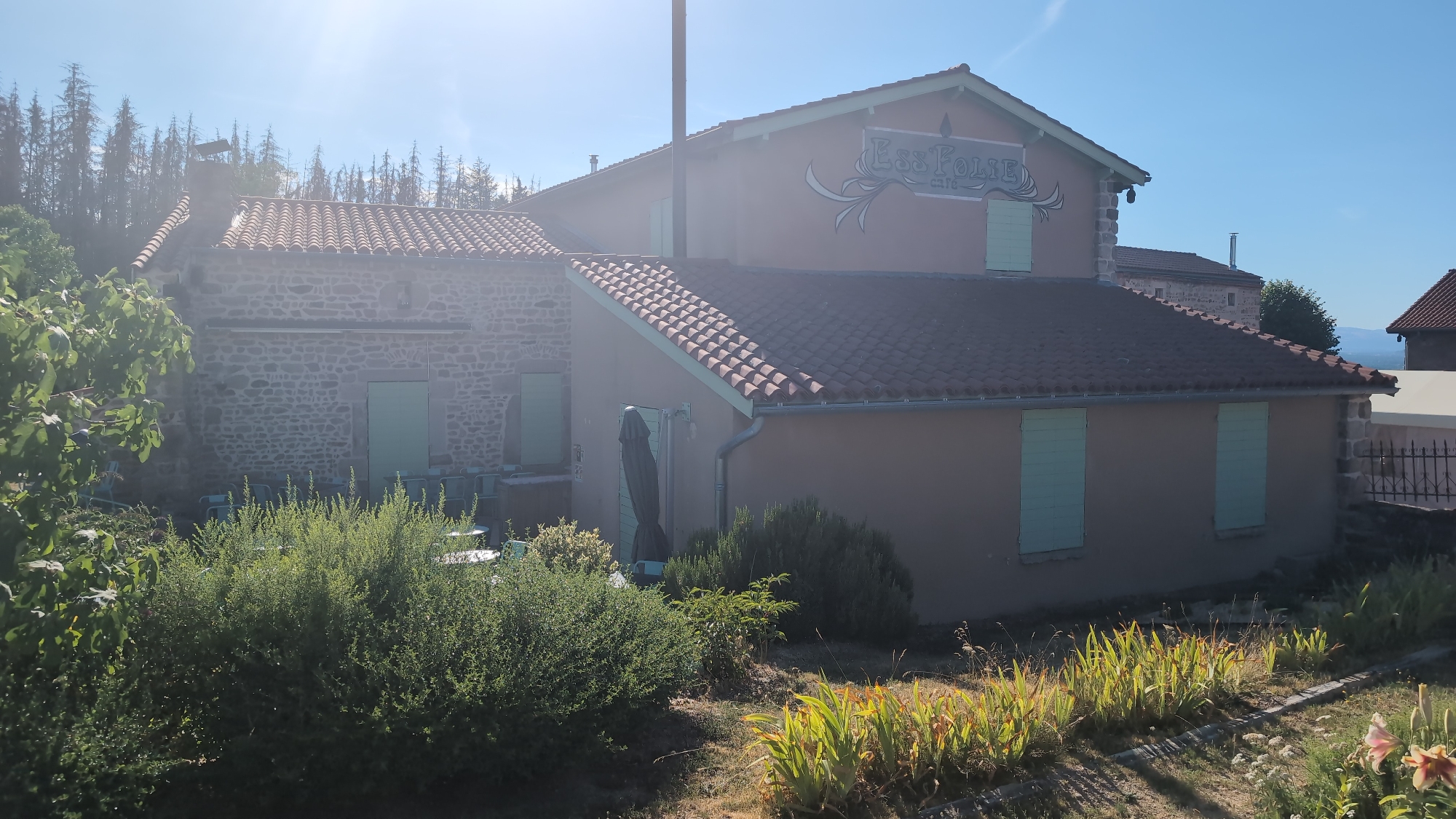 The image shows a building with a sign that reads "Ess'Folie Cafe" located in Chambles, France. The building has a terracotta tiled roof and is partially covered in stucco and stone. It has light green shutters on the windows. There is vegetation, including trees and bushes, in front of the building. The sky is clear and blue. The sun is shining brightly, creating a lens flare effect in the upper left corner of the image. The scene appears to be an outdoor seating area or a cafe setting.