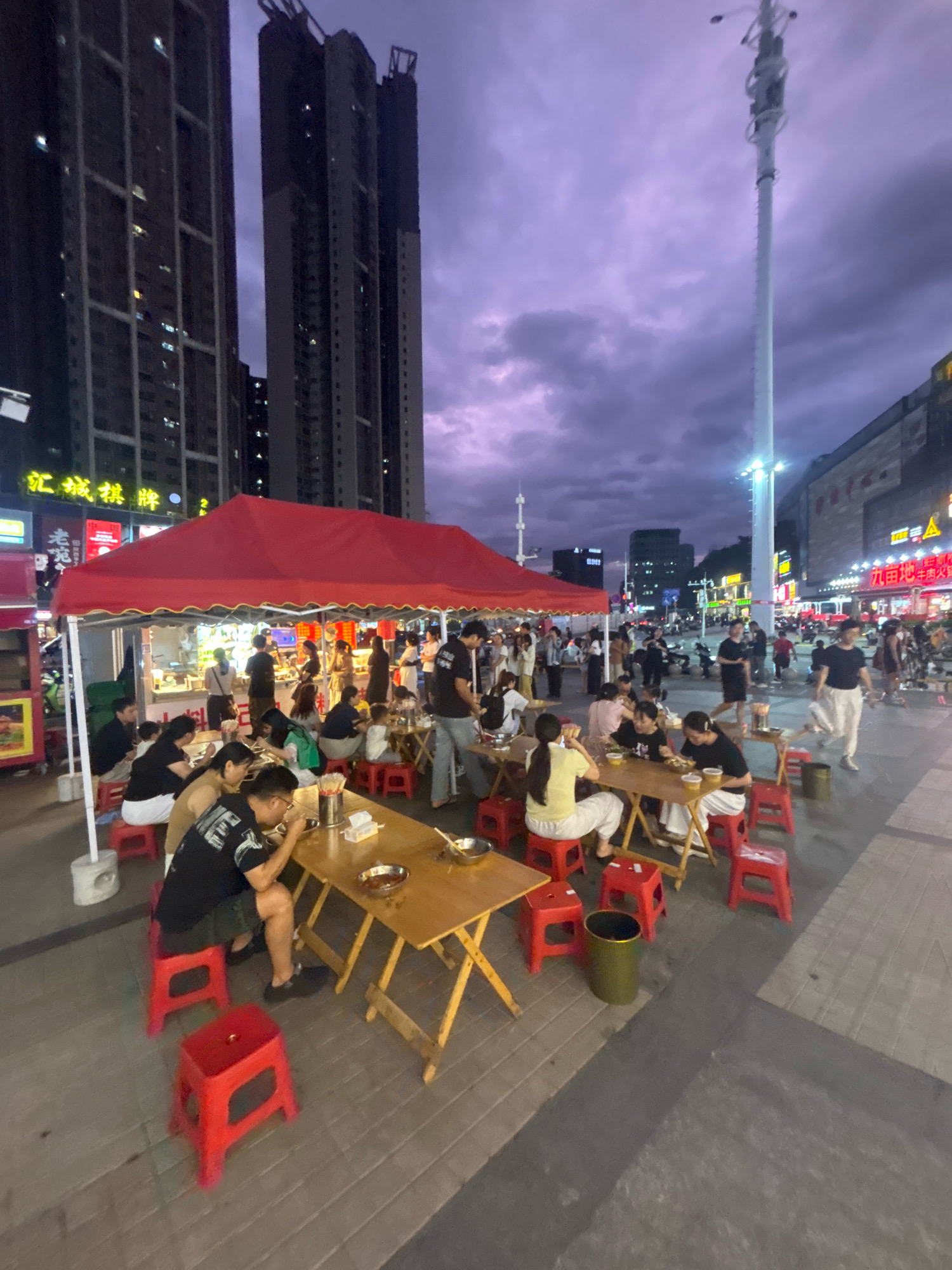 The image depicts an outdoor dining scene at dusk in Nanhai District, China. A red tent covers a section of tables and chairs where numerous people are seated, eating and conversing. The tables are set with food bowls, utensils, and drinks. Many individuals are casually dressed, and some are actively engaged in eating. Red plastic stools are arranged around the wooden tables. In the background, tall buildings and a street with a tall white tower and streetlights suggest an urban environment. The sky is a deep purple, indicative of twilight. The overall atmosphere is that of a lively, casual outdoor eatery.