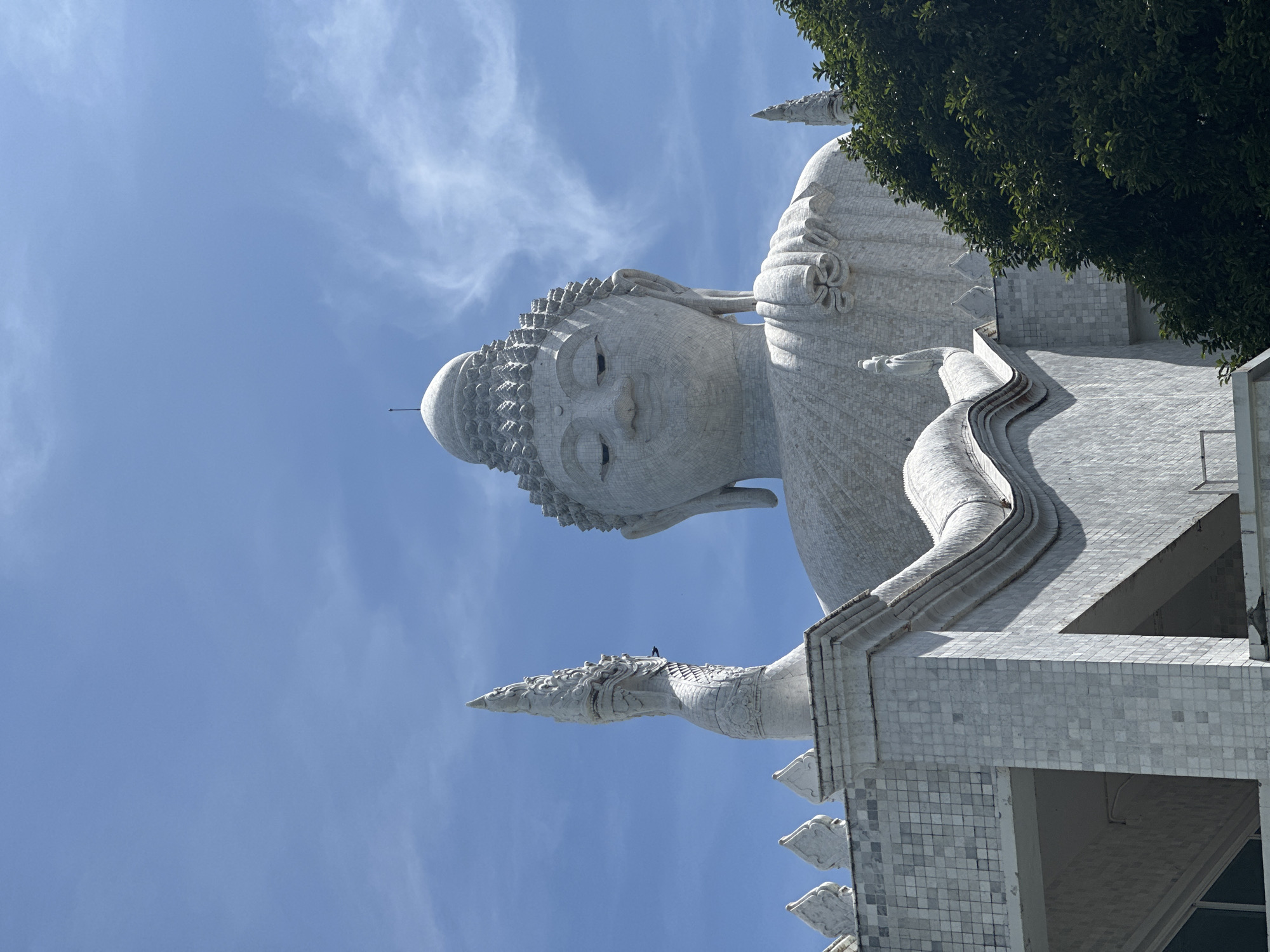An extreme low-angle, medium shot captures the monumental white marble statue of the Big Buddha against a bright blue sky with wispy white clouds. The statue dominates the frame, with its serene face and intricate details visible. In the foreground, a portion of the tiled base of the statue is visible, along with architectural elements adorned with white tilework and decorative spires. A sculpted Naga, a mythical serpent, rises to the left of the frame, its head pointed upwards. Lush green trees frame the right side of the image. The scene is static and appears to be a religious or tourist site, likely in Karon, Thailand, given the architectural style and the presence of a large Buddha statue. No people are visible in the frame.