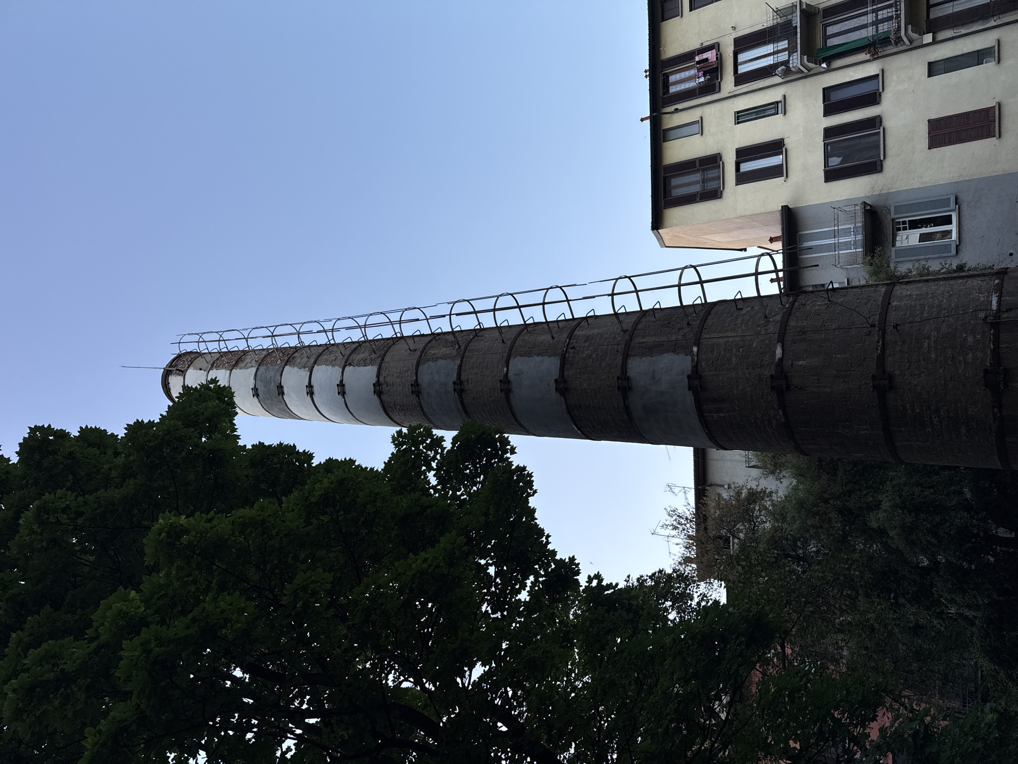 An upward-angled shot captures a tall, brick-like chimney or pipe extending horizontally from the side of a building. The chimney is segmented by metal bands and topped with a metal safety rail. The building is multi-storied with numerous windows, some covered by shutters. The lower portion of the image features a dark mass of tree foliage, partially obscuring the base of the chimney. The sky is a light blue, devoid of clouds. This is an industrial chimney in the city of Milan, Italy.