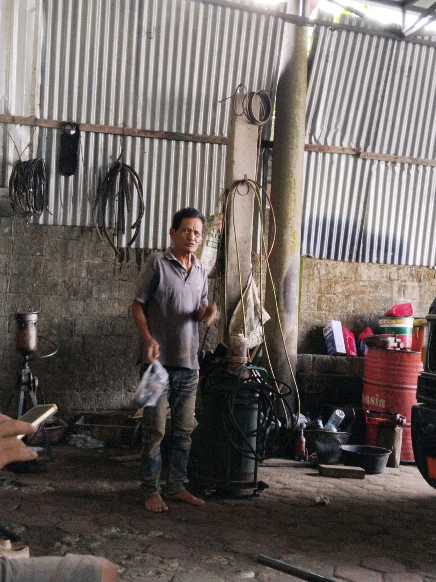 The image depicts a man standing in what appears to be a workshop or garage in Prigi, Indonesia. The man, of Asian descent, is wearing a grey polo shirt and denim jeans, and is barefoot. He is holding a plastic bag in his right hand and gesturing with his left. The environment is industrial, with a corrugated metal wall forming the background. Various objects are present, including coiled hoses, a large green tank, a red barrel, buckets, and boxes. The floor is concrete and appears to be dusty or dirty. The lighting suggests it is daytime, with natural light entering the space from above.