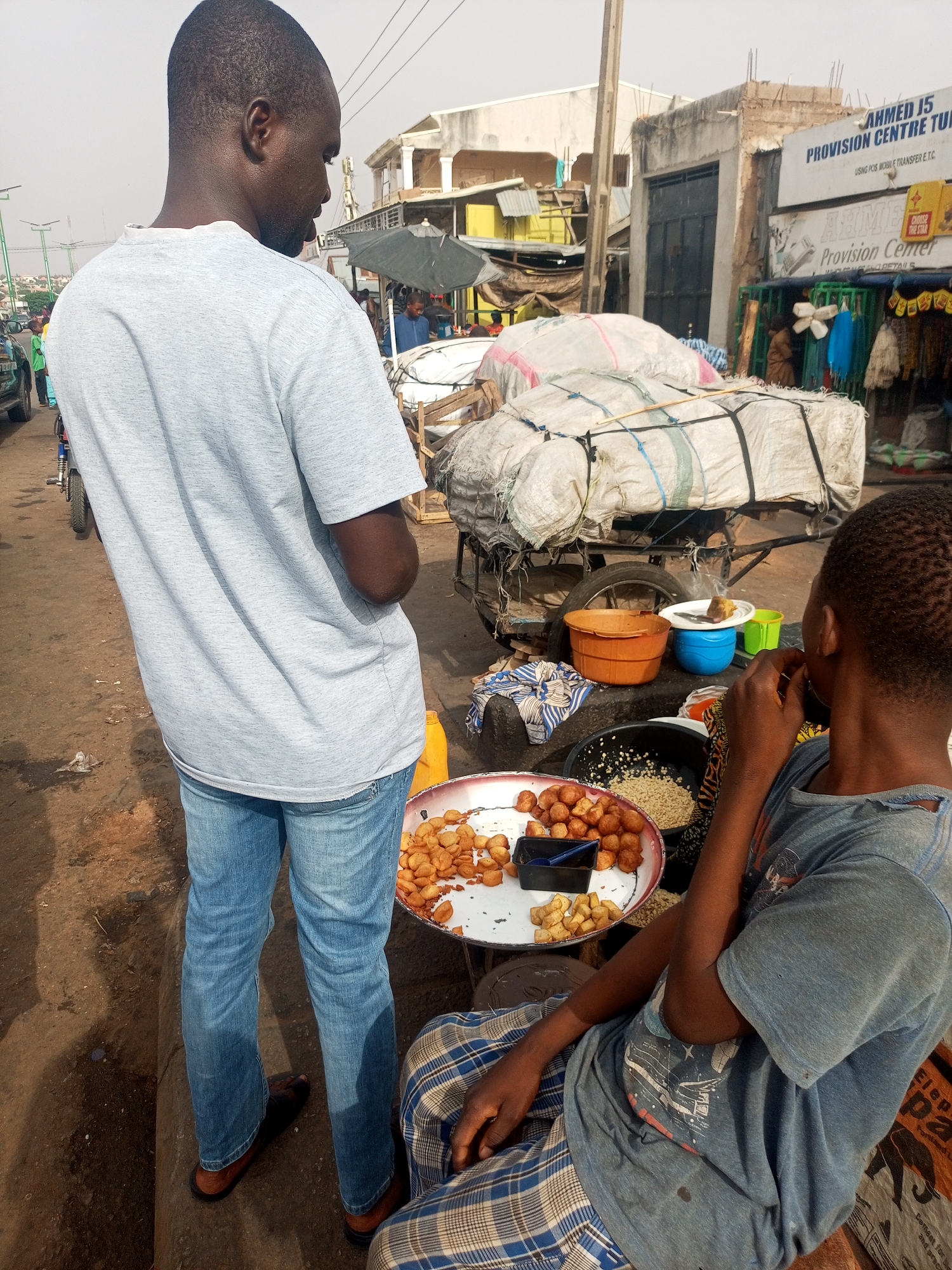 A man wearing a grey t-shirt and jeans stands over another man seated and eating snacks in what appears to be a street scene in Tempure, Nigeria. The seated man wears a blue t-shirt, and plaid pants and is sitting on a bag of Portland Cement. The street is filled with people and carts carrying goods, with businesses in the background that include the signs "Ahmed J5 PROVISION CENTRE TUN" and "THE STAR Provision Center". The surface of the street and the area around the men are visibly dirty.