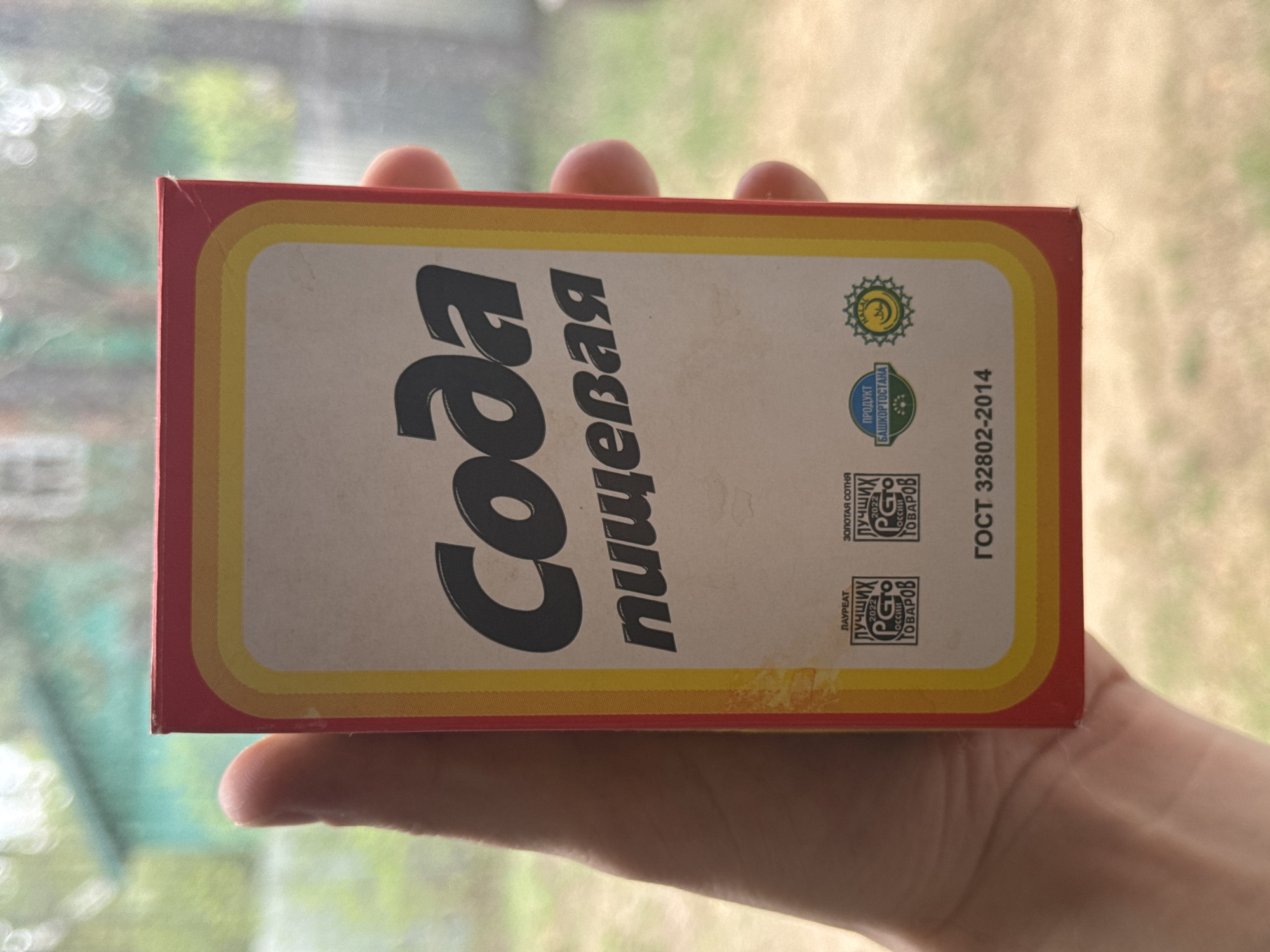 A hand holds a red and white cardboard box labeled "Soda" in Cyrillic. The box also has smaller logos and text indicating "Product of Bashkortostan" and compliance with GOST 32802-2014. The background shows a blurry outdoor scene with greenery, possibly taken near a window.