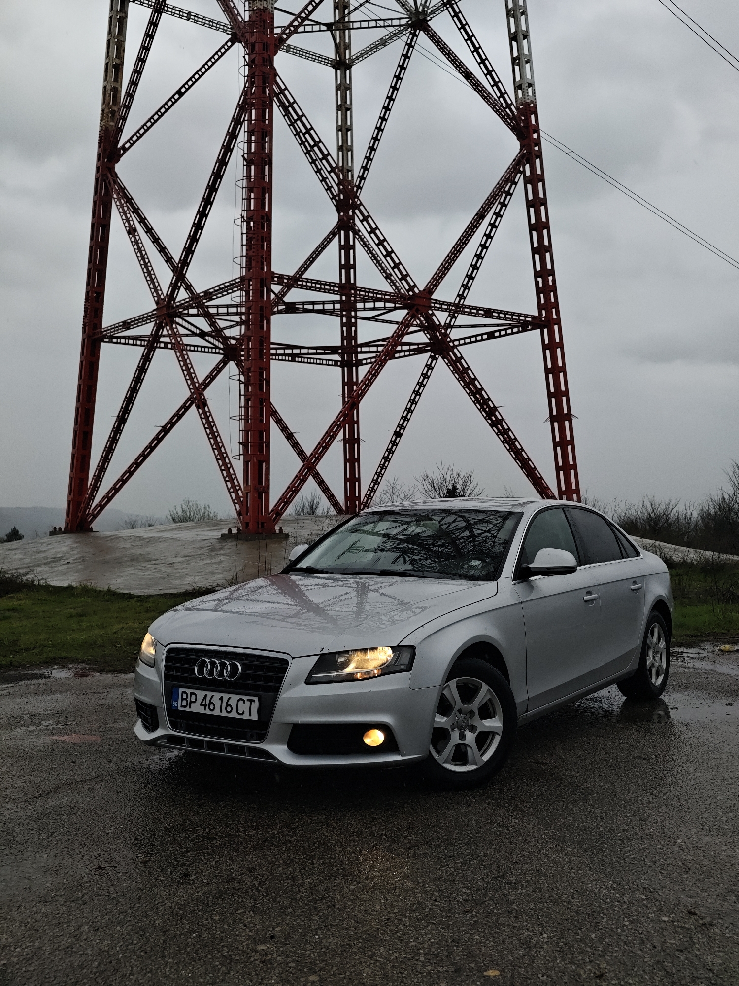A silver Audi A4 (B8 generation) sedan is depicted parked on a wet, dark gray aggregate surface under an overcast sky. The vehicle is positioned in a front-left quarter view with its headlights and fog lights illuminated. Water droplets are visible on its exterior, indicating damp conditions. A Bulgarian license plate, "BP 4616 CT," is affixed to the front bumper. In the background, a large, weathered, red-painted lattice steel power line pylon stands prominently. Its intricate structure features diagonal and horizontal bracing, with a ladder visible on one of its main vertical members. The pylon's base rests on a concrete foundation situated on an elevated, grassy, and somewhat barren terrain. Sparse, deciduous vegetation, devoid of leaves, is visible behind the car and surrounding the pylon's base. The scene is consistent with an area adjacent to a significant power transmission infrastructure, such as those found around the Kozloduy Nuclear Power Plant in Kozloduy, Bulgaria. No individuals or discernible actions are present.