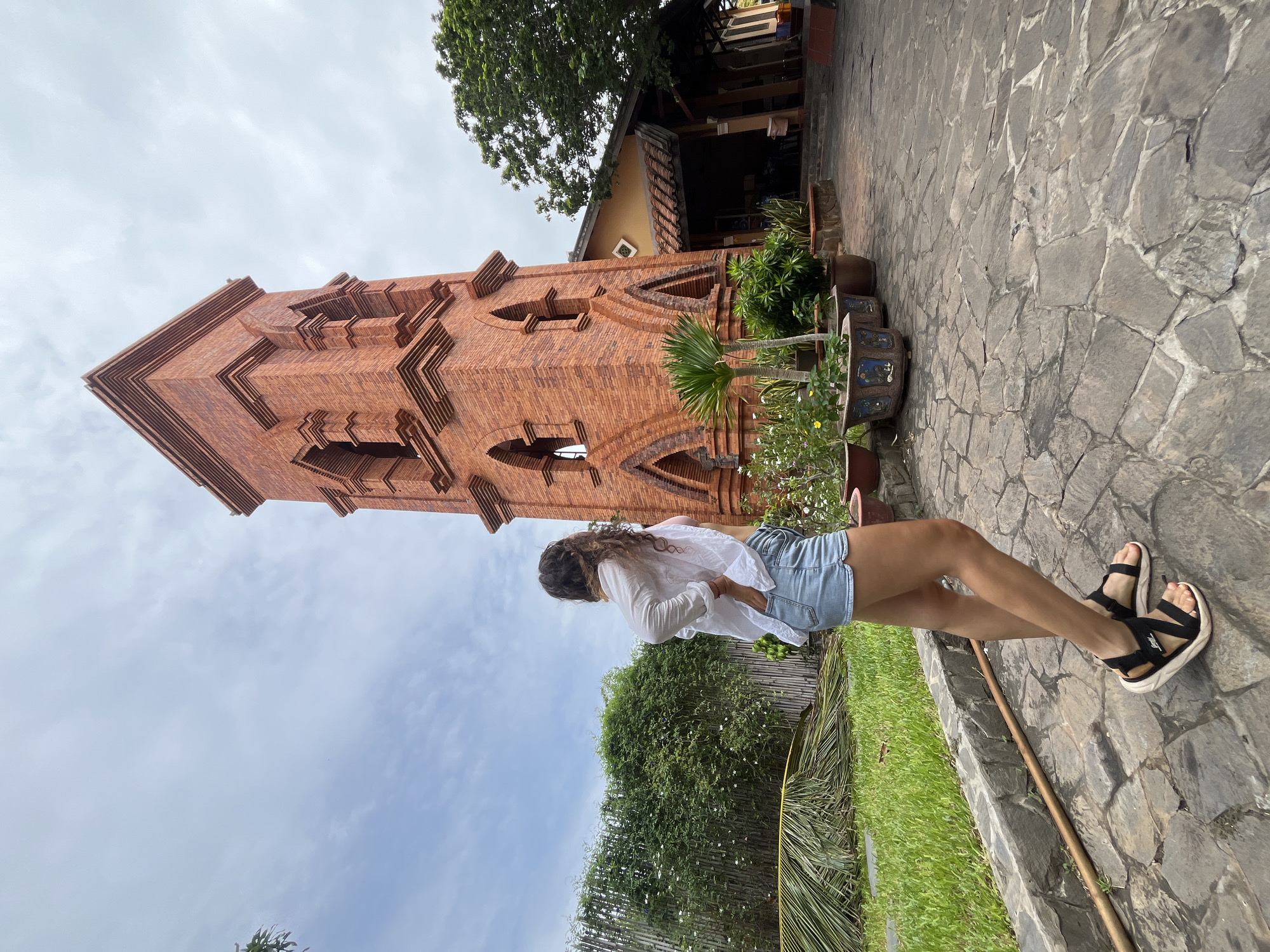 A young woman with curly brown hair, wearing a white open-neck shirt, light blue denim shorts, and black platform sandals, stands with her back to the camera in a courtyard. Her right hand is on her hip, and her left hand is in her pocket. She is looking towards a tall, red brick tower with arched openings and decorative brickwork, which is situated to her right. In front of the tower are several potted plants. To the left of the woman is a lush green lawn with a fallen palm frond. Behind the lawn, a bamboo fence is visible, covered in greenery and small white flowers. The ground in the courtyard is paved with irregularly shaped stone tiles. The sky above is cloudy with patches of blue. The scene appears to be set in the city of Phu Thuy Ward, Vietnam.