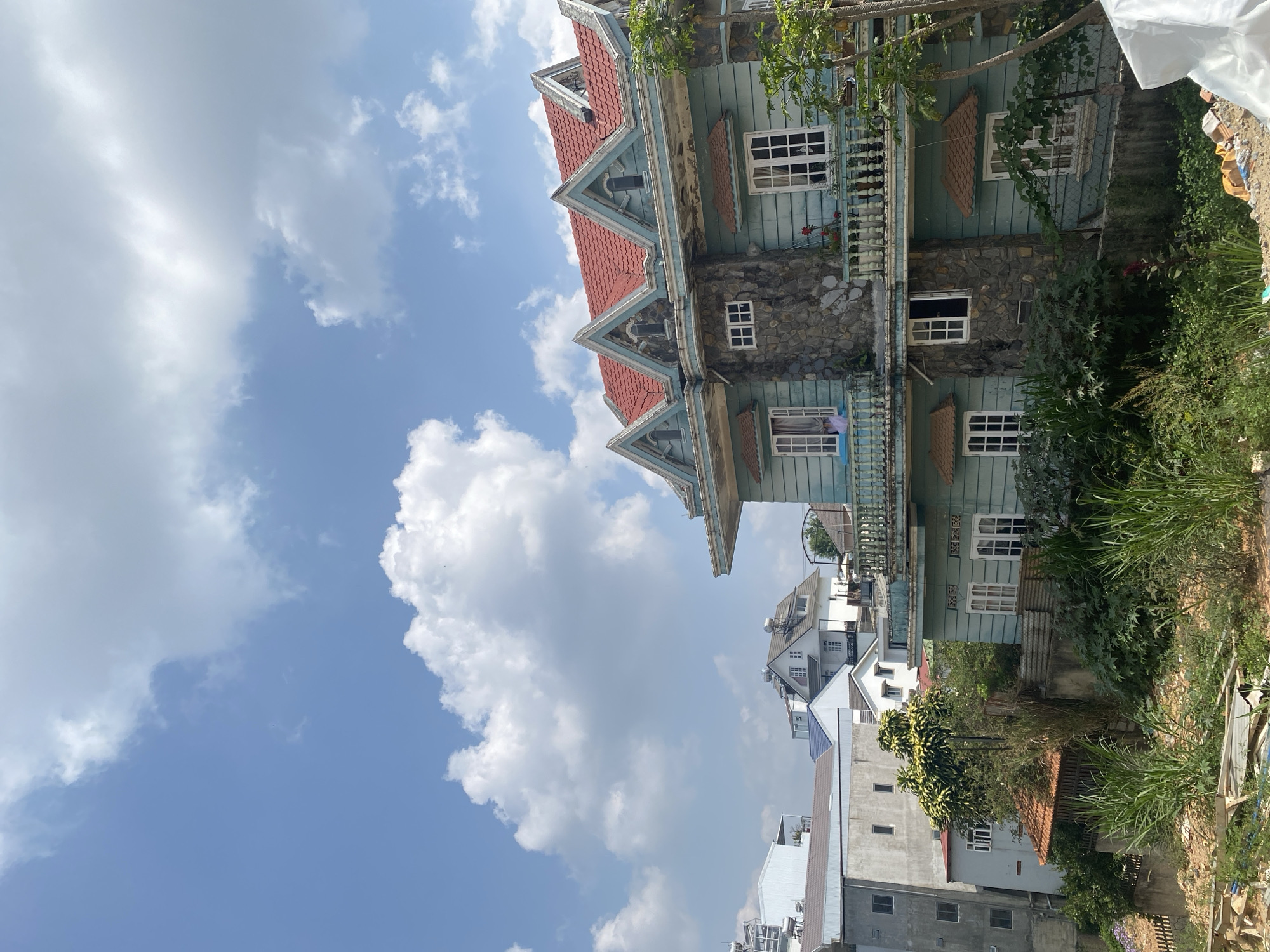 The image displays a multi-story house with distinct architectural features under a bright blue sky filled with fluffy white clouds. The house, positioned prominently in the midground, has a steep, red-tiled roof with multiple gables and decorative trim. Its upper levels feature light blue wooden siding, while the lower level is constructed from rough-hewn stone. Numerous white-framed windows are visible across both stories, and a white balustrade lines an upper balcony.
To the right of the house, lush green vegetation, including tall grasses and bushes, fills the foreground and midground. Some trees are also visible, partially obscuring parts of the house. In the background, several other buildings can be seen, appearing to be a mix of modern and traditional residential structures, some with white walls and dark roofs.
The scene depicts a sunny day, indicated by the clear blue sky and bright lighting, likely late morning or early afternoon. There are no people or specific activities visible. No text is discernible in the image. The architectural style of the main house and the surrounding greenery suggest a setting in a mountainous or hilly region, consistent with the provided location context of Lâm Viên - Đà Lạt, Vietnam.