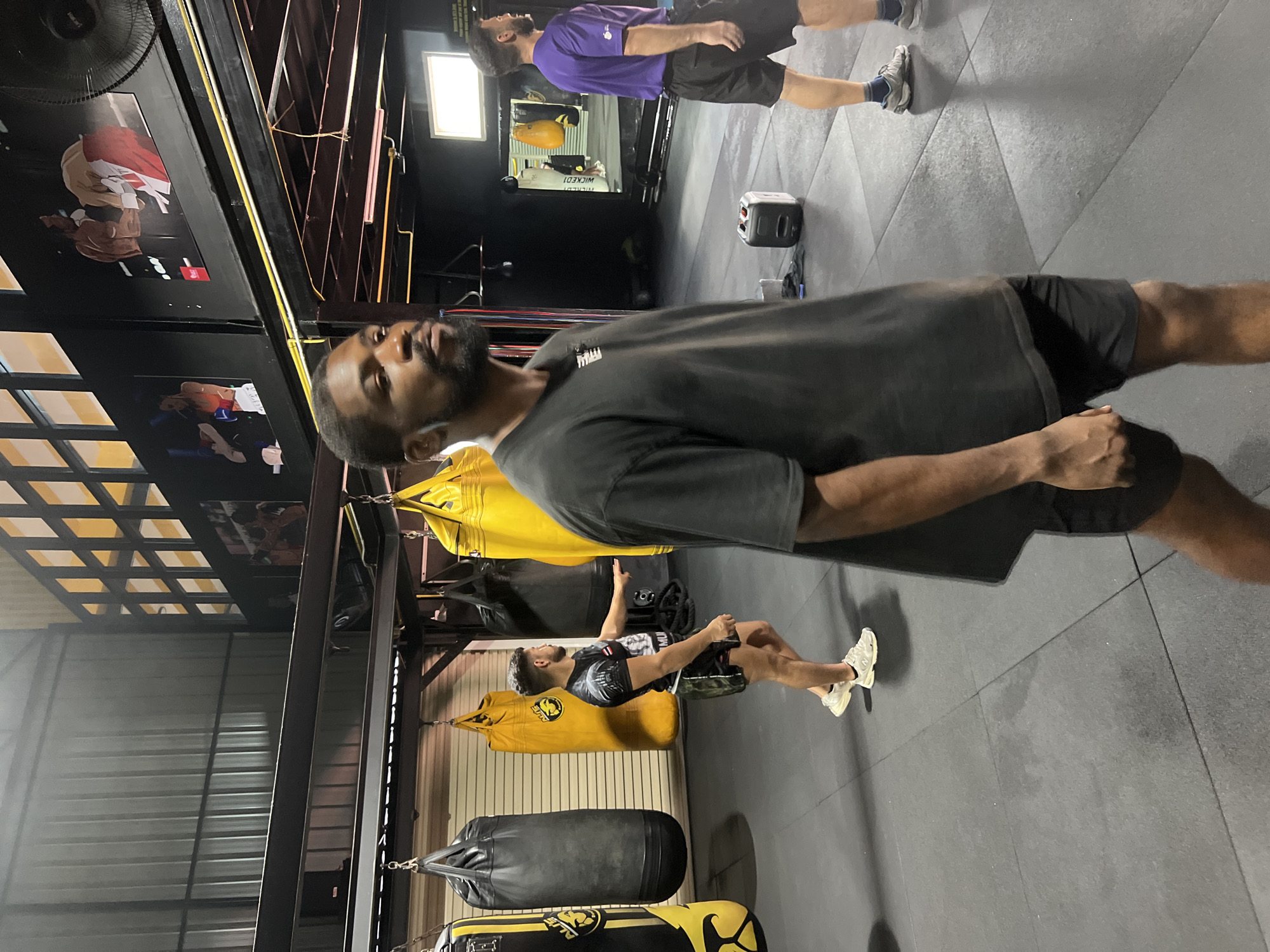 In a boxing gym located in Chalong, Thailand, a Black man with a beard stands in the foreground, facing right and looking upwards. He is wearing a dark grey t-shirt and black shorts. In the middle ground, a man with dark hair and a beard, wearing a black t-shirt with a graphic and camouflage shorts, points towards a punching bag. To his left, a large black punching bag hangs, and further to the left, a yellow punching bag is visible. Behind them, more punching bags and gym equipment are present. In the background, a man in a purple t-shirt and black shorts is partially visible, facing away from the camera. The gym has a dark grey mat floor, black metal structures, and motivational posters on the wall featuring boxers. Natural light seems to be coming from the left side of the image.