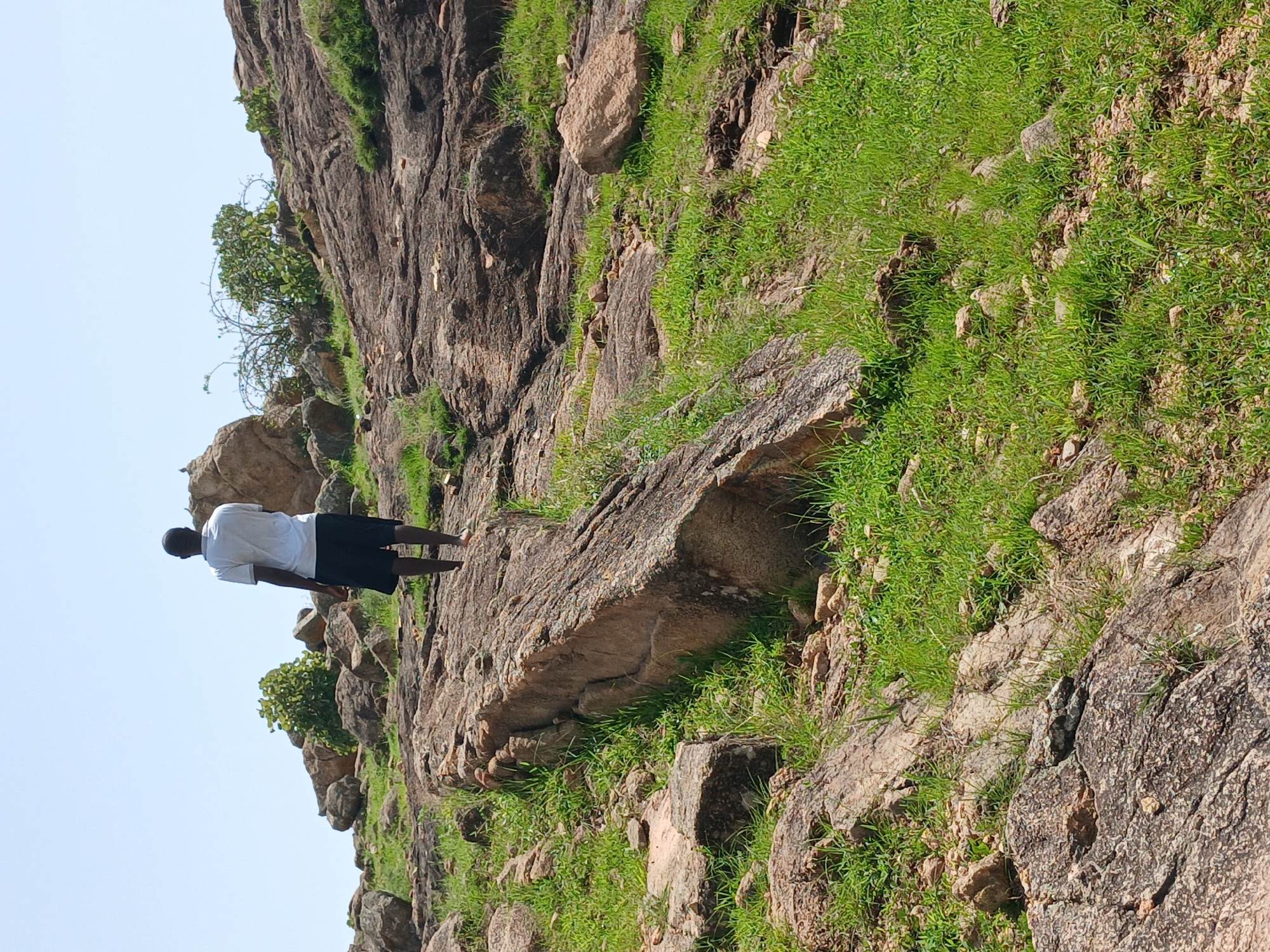 A person walks up a rocky, grassy hillside in Kwang, Nigeria. The individual is viewed from behind and is wearing a white t-shirt and dark shorts. The terrain is characterized by large, exposed rocks and patches of green grass. The sky is clear and pale blue.