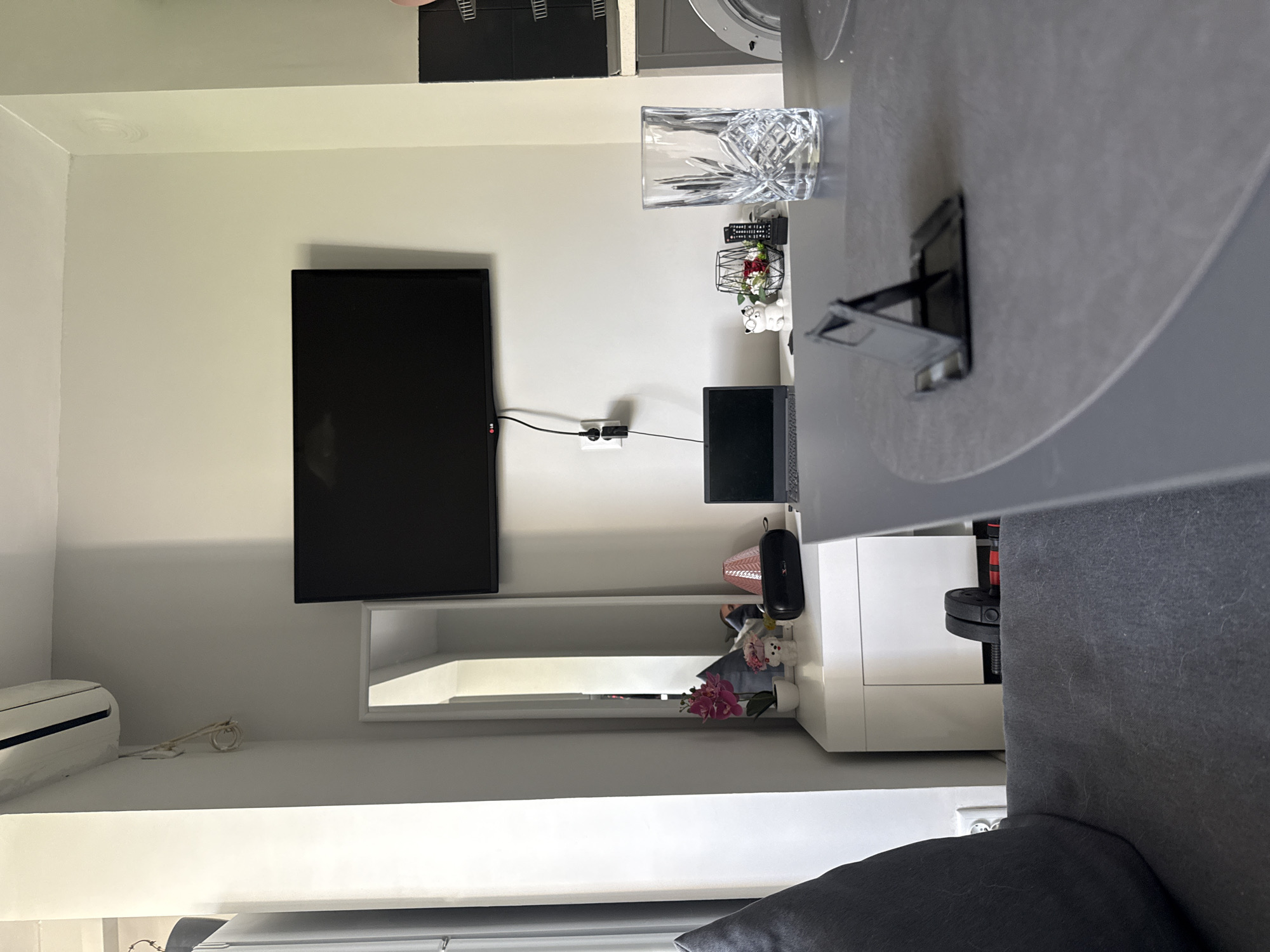 The image depicts an interior space. A television screen is mounted on a white wall above a white storage unit, which is partially visible below the screen. A laptop and various decorative items are placed on the unit. A crystal glass is on a table to the right of the screen. A gray chair occupies a portion of the foreground. An air conditioner is mounted high on a wall in the lower left corner. The location is not specified, but is presumed to be somewhere in Stara Zagora, Bulgaria.