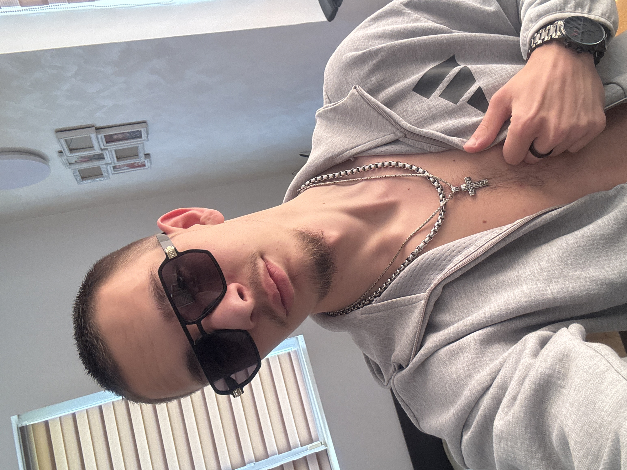 The image shows a man wearing sunglasses, a grey hoodie with an Adidas logo, and two silver necklaces with a cross pendant. He is also wearing a watch and a ring. He is partially unzipping his hoodie. The location appears to be indoors with blinds visible in the background, and a ceiling light fixture with multiple frames on the ceiling. There is no indication of the location being Vetren Dol, Bulgaria.