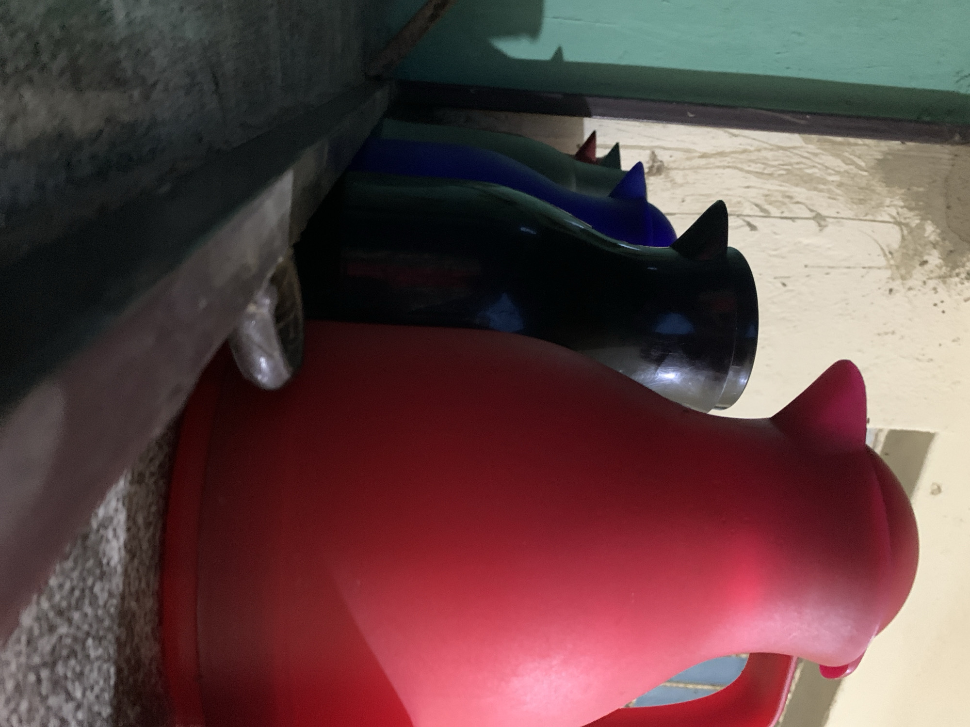 The image displays an indoor setting in New Stadium, Nigeria, featuring a low-angle perspective of a row of thermal beverage jugs positioned on a textured, greyish surface. Dominating the foreground on the right is a large, matte red thermal jug, characterized by its rounded form, a distinct spout, and a partially visible handle. To its immediate left and slightly behind, a black thermal jug is visible, followed by a blue jug, a green jug, and a final jug with a red-colored spout, each partially obscured by the preceding item. A dark, linear element, possibly a metal beam or wooden edge, extends diagonally across the lower portion of the frame from bottom-left to mid-right. A small, reflective, metallic object rests upon this beam. The background consists of a light-colored wall with multiple irregular dark streaks and smudges. A vertical section of a teal or light green wall or frame is present on the far left, separated by a darker vertical trim. A structural outline, consistent with a window or opening, is faintly discernible in the upper-right background. No individuals or dynamic actions are depicted.
