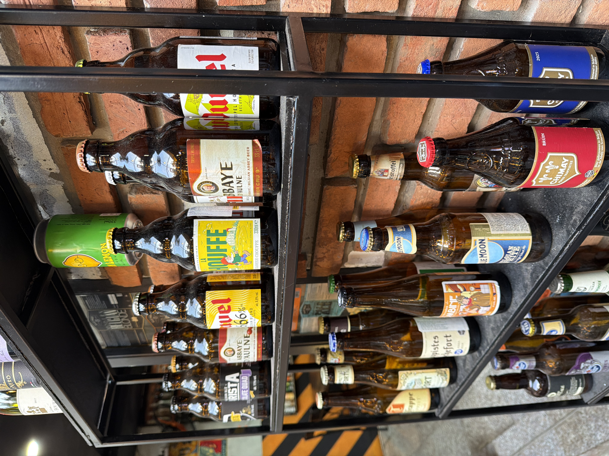 The image displays a shelf of bottled beers, predominantly Belgian brands, arranged on a black metal shelf against a brick wall. The bottles vary in size and label design, with some featuring prominent brand names like "La Chouffe," "Duvel," and "Chimay." A single green can, possibly a beverage, is visible on the top shelf. The shelf is part of a display in what appears to be a retail or bar setting. The context suggests the location might be a specialty beer store or a restaurant in South Quy Nhon Ward, Vietnam, where such international beverages are often sold. There are no people visible in the image.