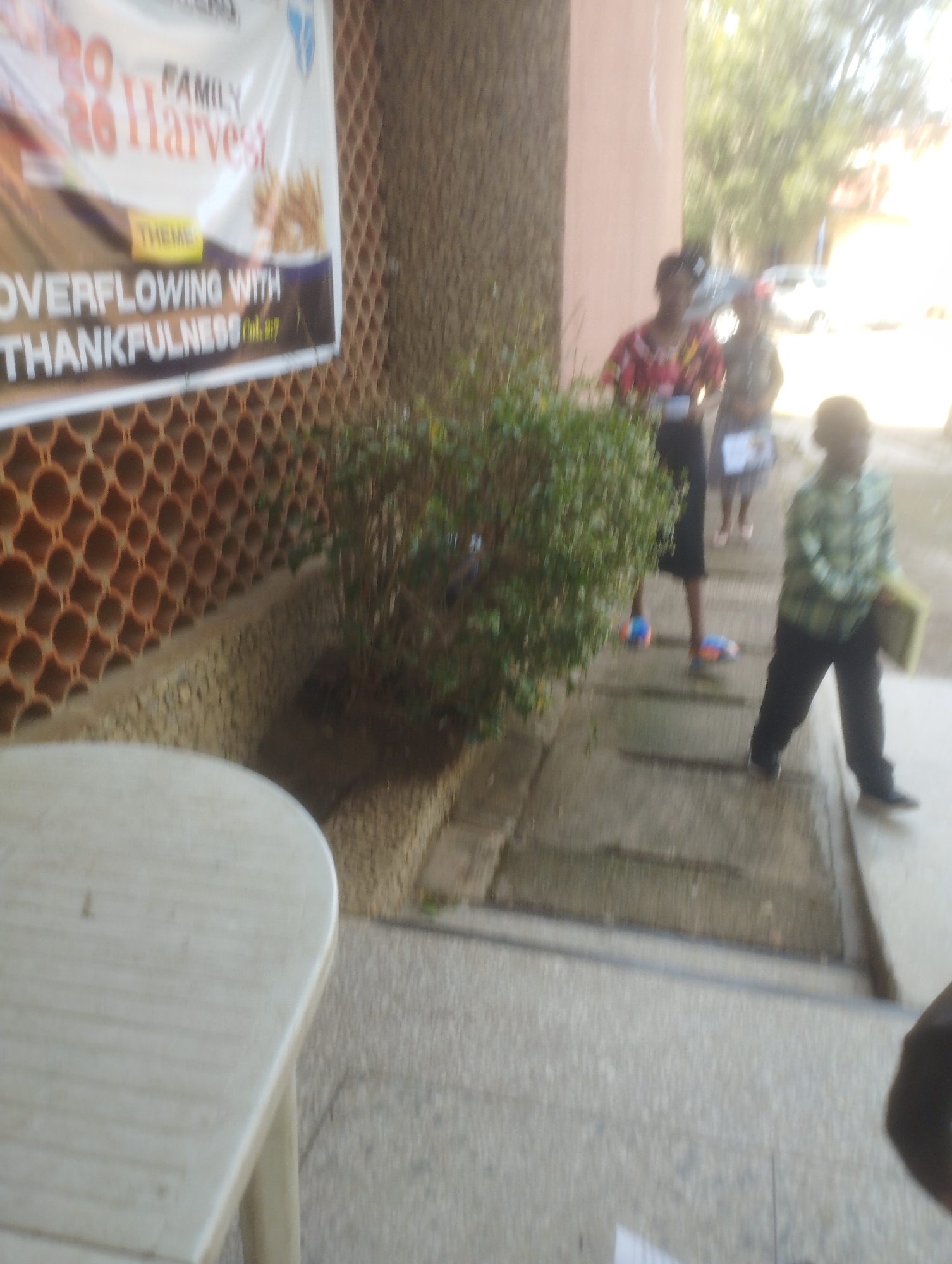 The image shows a banner with text that includes "2020 FAMILY HARVEST" and "OVERFLOWING WITH THANKFULNESS Col 3:17". There are several people in the background, including two individuals wearing patterned shirts and one person in a checkered shirt. The setting appears to be outdoors, with a wall constructed of cylindrical bricks on the left and a tree or bush in the middle ground. A light-colored table is visible in the foreground on the left. The scene is blurry and overexposed in some areas, particularly in the upper right. There is no specific information in the image to indicate it is located in Jos, Nigeria.