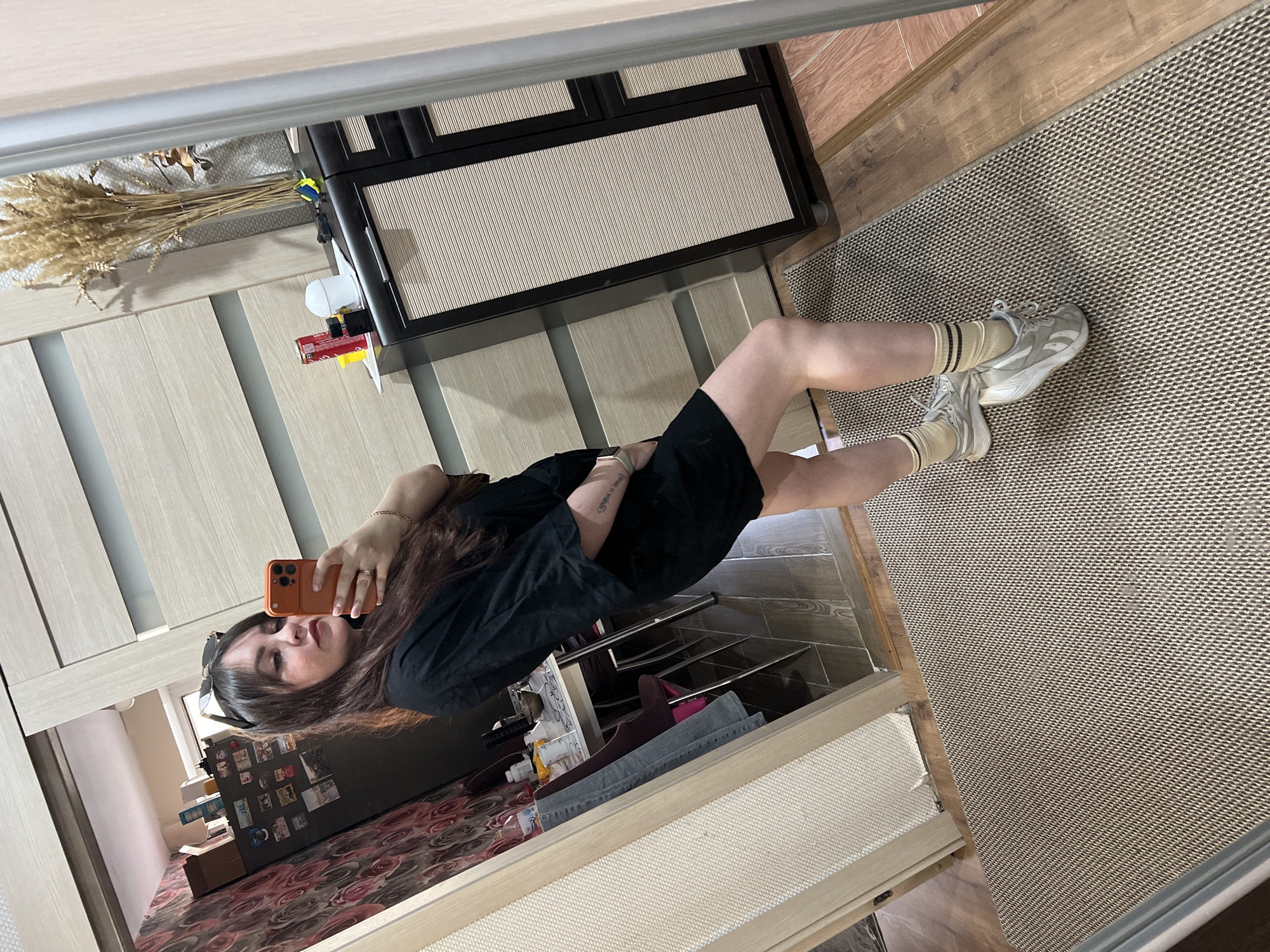 A woman is taking a selfie in a mirror. She is wearing a black shirt, tan socks with black stripes, and white sneakers. The background includes a dresser with black and beige drawers, a closet with beige doors, and a rug with a geometric pattern. There are also decorative items such as dried wheat and various objects on shelves in the reflection. The image doesn't contain enough data to infer the location in the city of Noviy, Russia.