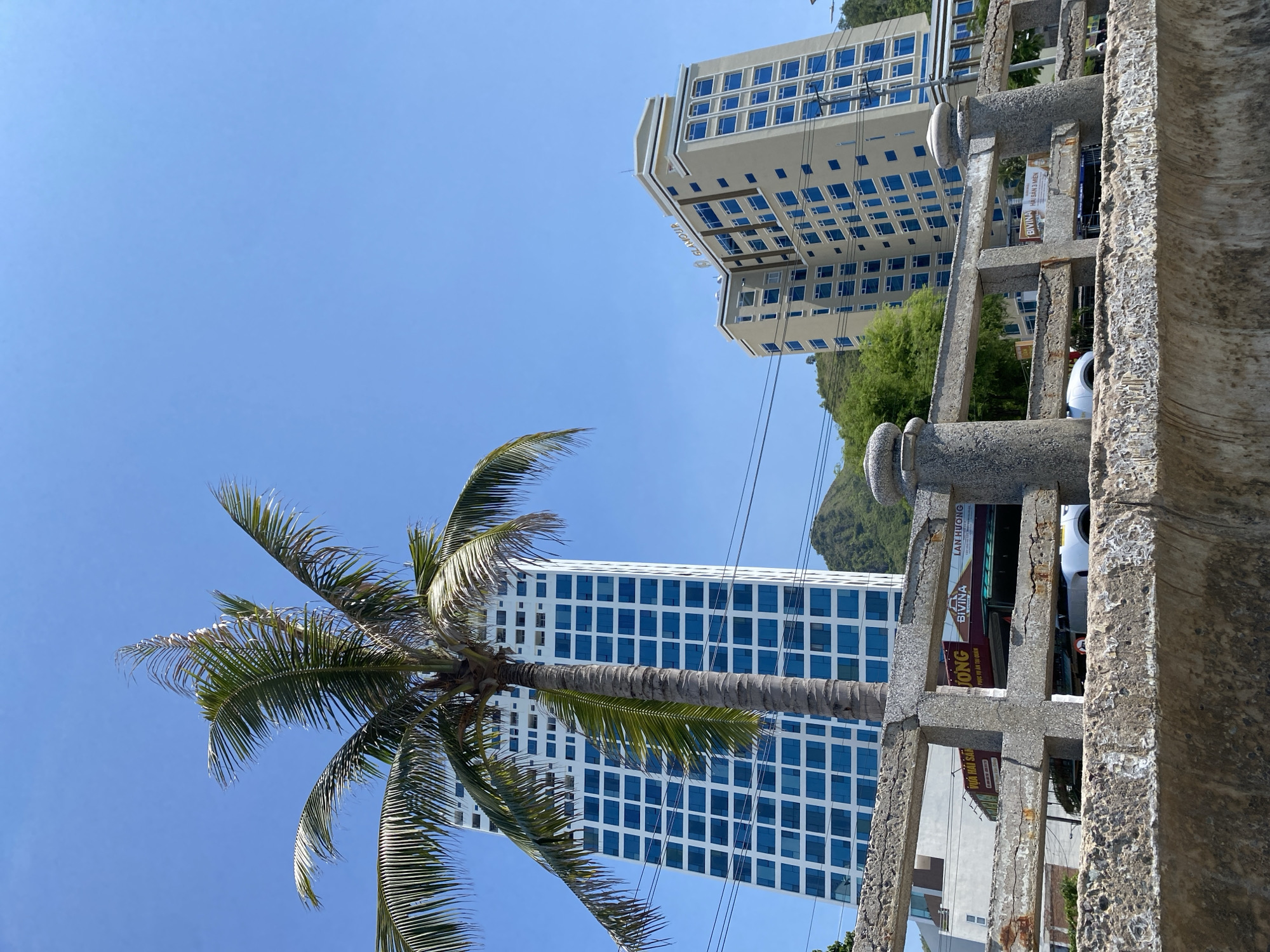 A bright, sunny day in Bac Nha Trang Ward, Vietnam, is captured in this low-angle shot looking up at modern buildings and a palm tree. The foreground is dominated by a weathered, concrete railing with a rough texture and visible cracks, supported by cylindrical pillars. Beyond the railing, a tall, slender skyscraper with a white facade and numerous blue-tiled windows stands prominently, partially obscured by a mature palm tree with vibrant green fronds. To the right, a tan-colored, multi-story building with a similar pattern of blue windows and a sign that reads "GLAMOUR" on its top level is visible. In the background, lush green hills rise beneath a clear, brilliant blue sky. The scene suggests a coastal or resort town environment, with the architecture and vegetation indicative of a tropical location. There are no people clearly visible in the image, but the presence of cars and businesses suggests a populated area.