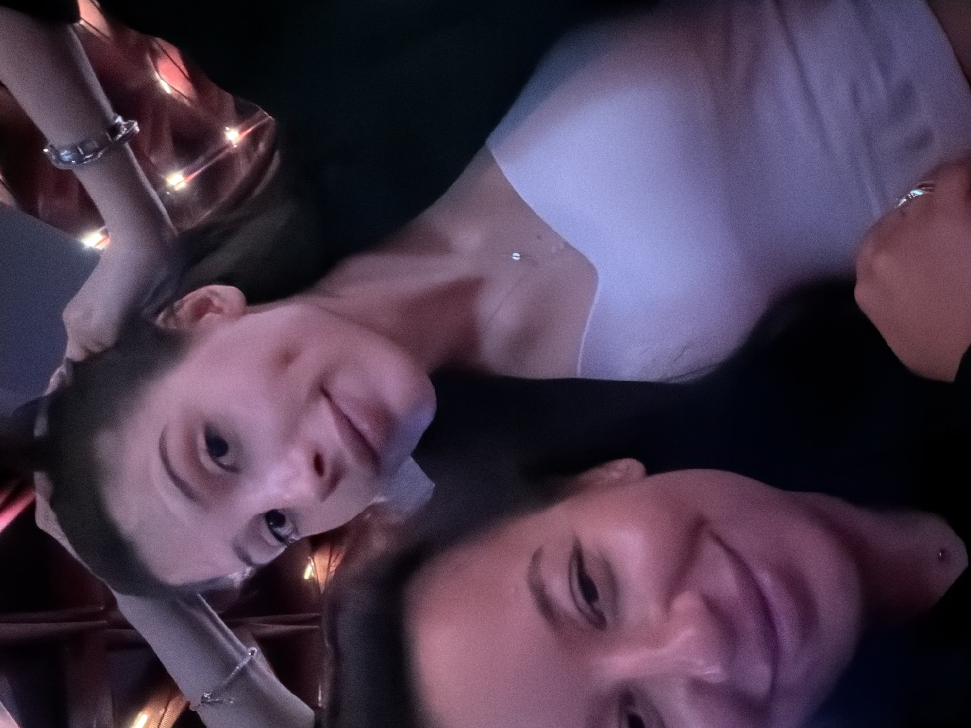 Two women are taking a selfie. The woman in the foreground, on the left, is smiling and looking directly at the camera. She has dark hair and is wearing a black top. The woman in the background, on the right, is also looking at the camera. She has light brown hair and is wearing a white top and a black jacket. Her hands are in her hair. There are string lights in the background, creating a warm, festive atmosphere. The lighting suggests this photo was taken indoors, possibly at night, in a dimly lit environment with decorative lighting. The overall mood of the image is casual and friendly.