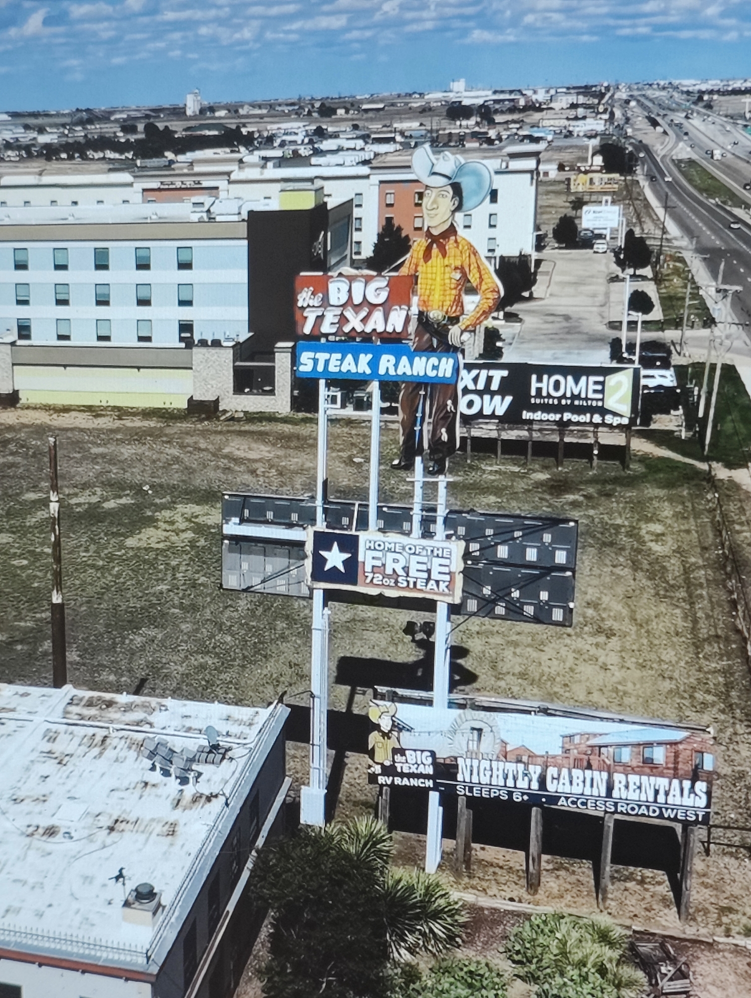 The image is an aerial view of a commercial area, likely a roadside attraction and lodging, under a cloudy sky. Dominating the scene is a large sign for "The Big Texan Steak Ranch," featuring a cartoon cowboy. Adjacent to this are signs for "Home 2 Suites by Hilton" and "Nightly Cabin Rentals." In the background, a highway with traffic is visible, alongside various buildings that appear to be hotels and businesses. A rooftop view of one building shows what appears to be HVAC units or other rooftop equipment. The ground is mostly undeveloped, with patches of grass and dirt. The location is not Yarmolyntsi, Ukraine.