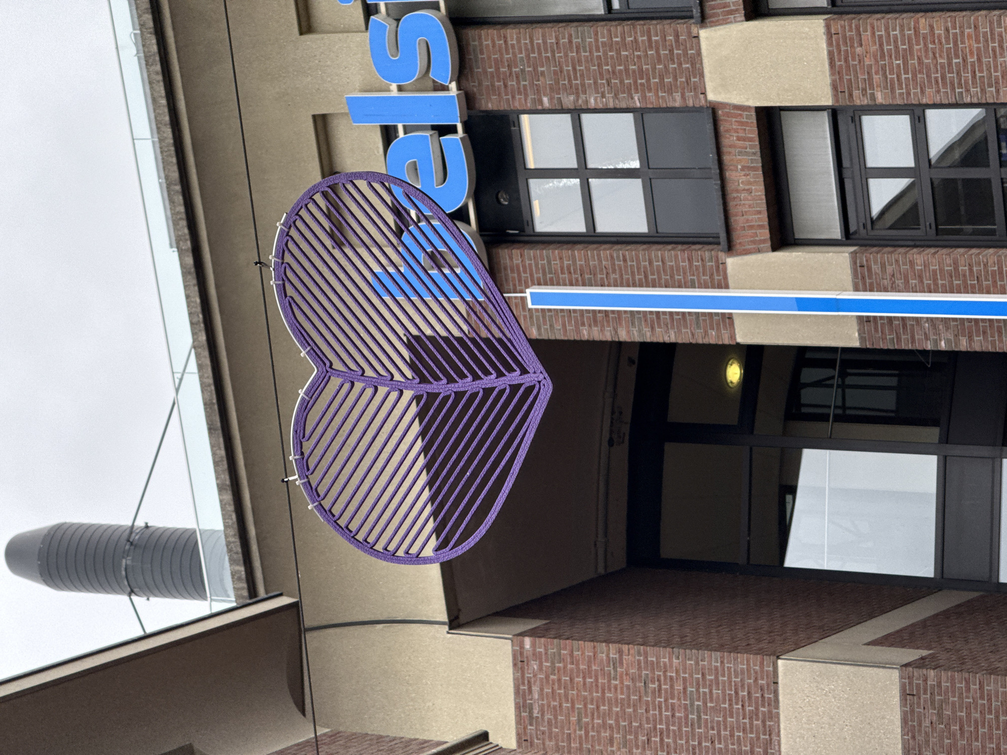 The image shows the exterior of a building with a prominent purple heart-shaped art installation hanging in front of it. The building's facade is a mix of brickwork and lighter-colored sections. Large blue letters spelling out "belsi" are visible on the upper part of the building, suggesting it's a commercial establishment. Several windows with dark frames are present on the building's exterior. A tall, dark gray cylindrical structure, possibly a chimney or ventilation shaft, is visible in the background. A thin, blue vertical sign is also attached to the building. The sky is overcast. The scene appears to be located on a street in the city of Groningen, Netherlands, as indicated by the "Belsi" signage, which is associated with a Dutch insurance company. There are no people visible in the image.