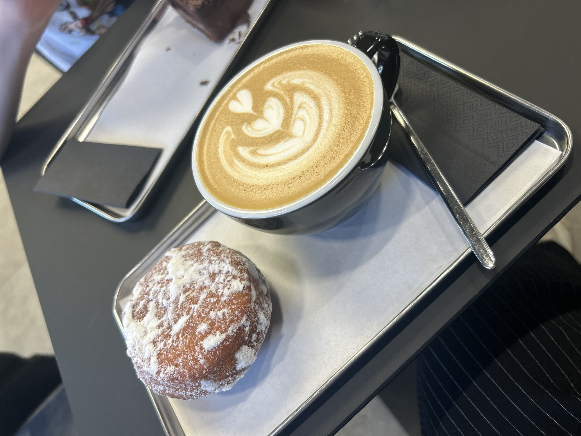 The image depicts a cup of coffee with latte art and a powdered donut on a metal tray. The tray also contains a spoon and a black napkin. A second tray with a brownie is partially visible in the upper left corner. The background is a dark, possibly black table. A person wearing pinstripe pants is partially visible in the lower right corner. The location in the city of Izhevsk, Russia is unknown.