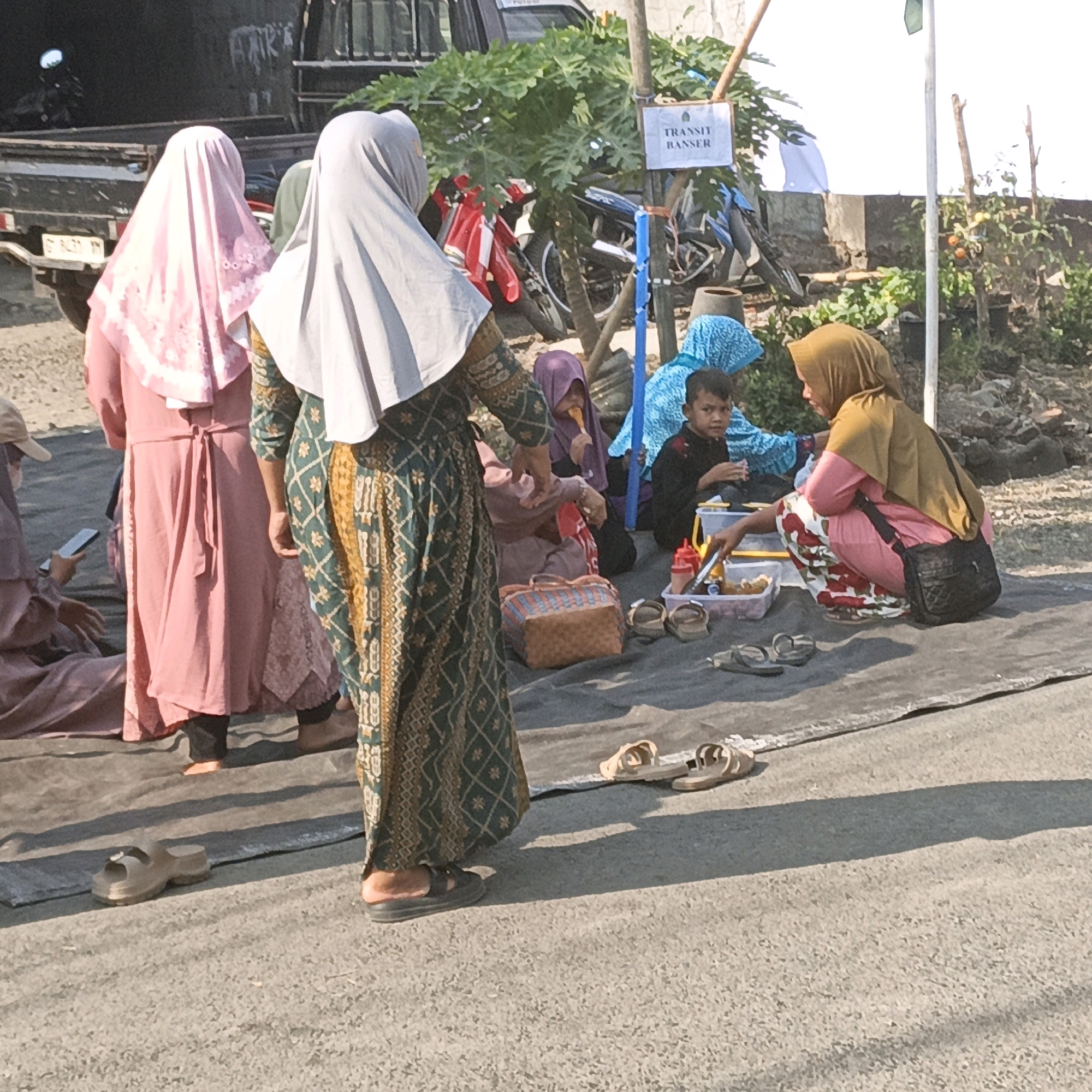 In the city of Kalitorong, Indonesia, a group of people, mostly women and children, are gathered outdoors. Several individuals are wearing traditional Islamic clothing, including headscarves (hijabs) and long dresses. Some are seated on a dark tarp laid out on a paved surface, while others are standing. In the background, a truck is parked, and motorcycles are visible, suggesting a street or roadside setting. A sign reading "TRANSIT BANSER" is displayed on a pole. There are containers with food items and utensils, indicating a potential gathering or event. The environment appears to be sunny, with some vegetation and a wall in the background.