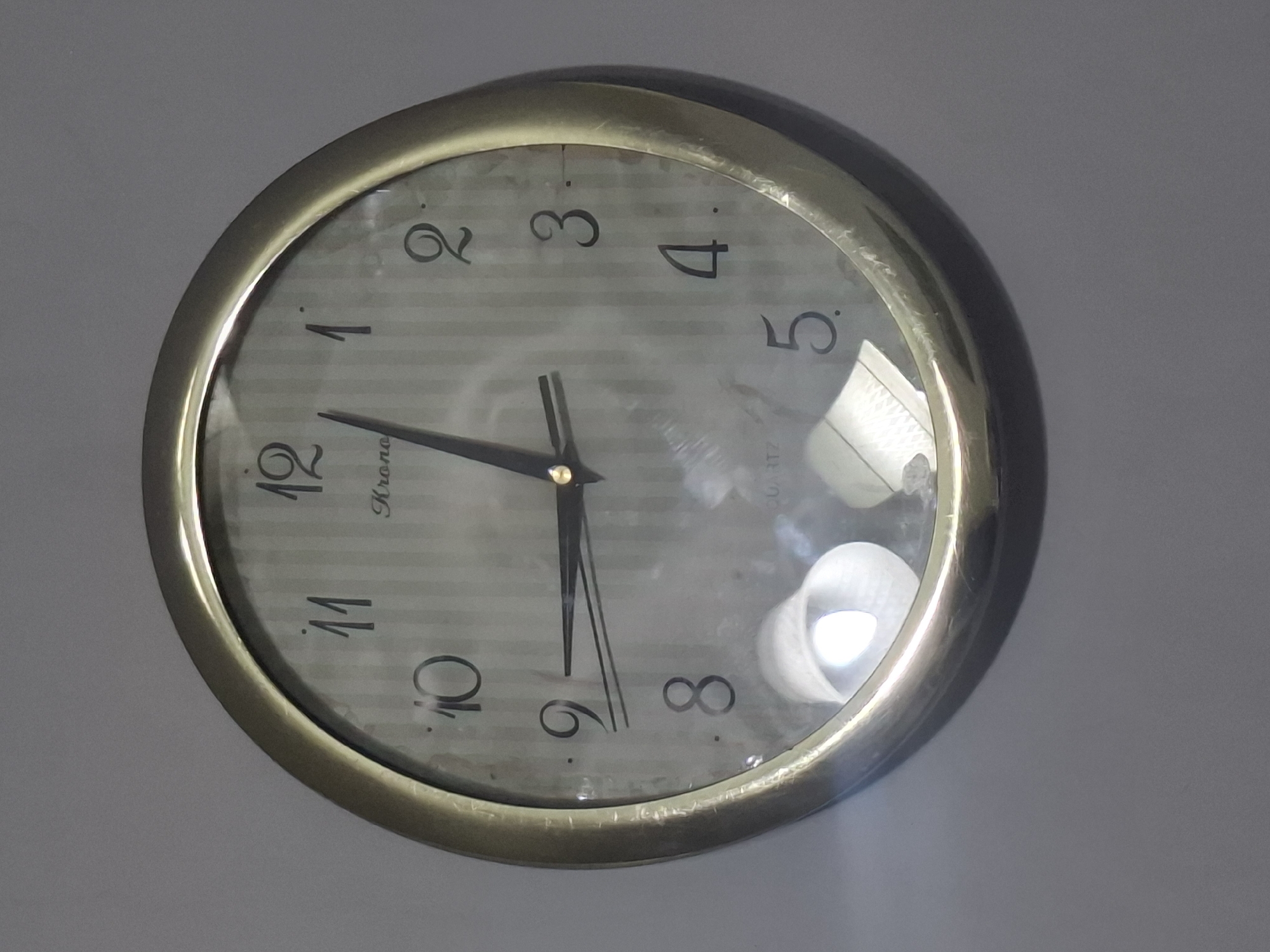 A round, analog wall clock with a metallic frame is mounted on a plain, gray wall. The clock face has a striped pattern and displays black Arabic numerals for hours. There are three hands: a short hour hand, a long minute hand, and a thin second hand, all black. The hour hand is pointing slightly past the 12, the minute hand is pointing at the 2, and the second hand is near the 12. The word "Kronos" is visible on the clock face, along with "Quartz" at the bottom. There are reflections on the clock's glass surface, including what appears to be a bright light source and possibly some textured material below the clock. The image does not contain any information or visual cues that would allow for the identification of its location in the city of Almaty, Kazakhstan.