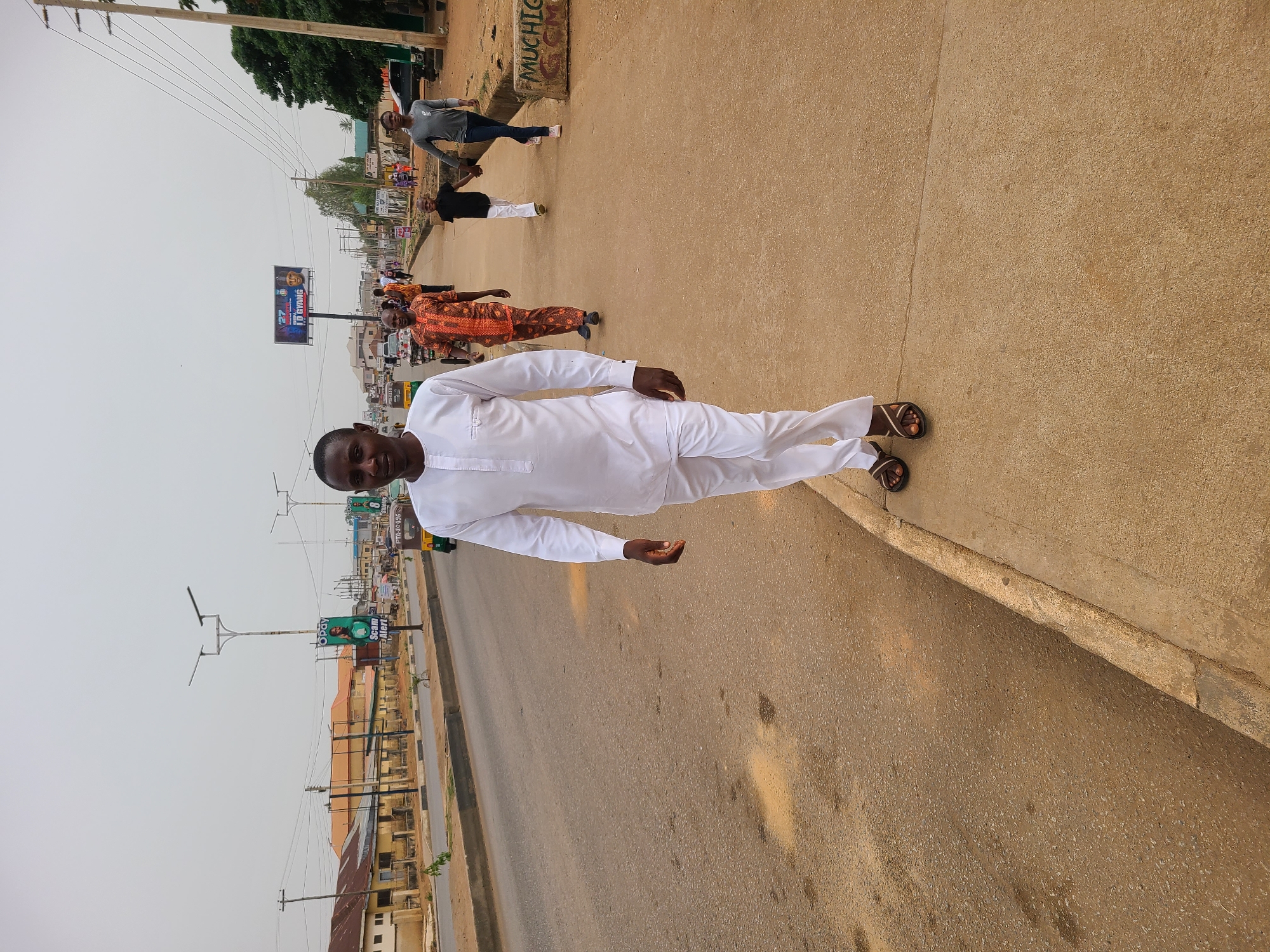 The image captures a full-body view of a man standing on a sidewalk in what appears to be the city of Jos, Nigeria. He is wearing a white long-sleeved outfit and brown sandals. In the background, there's a street scene with other pedestrians, buildings, streetlights, and billboards visible. Overhead power lines are also present. The sky appears overcast. A road is running adjacent to the pavement the man is standing on.