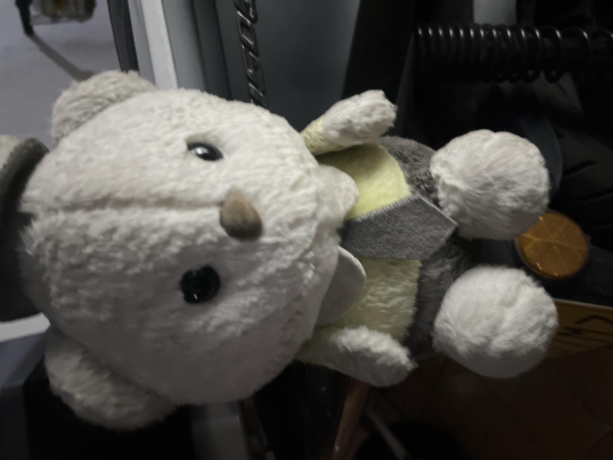 A white teddy bear is seated and facing the camera. The bear is wearing a yellow shirt with a gray tie. It has black button eyes and a brown nose. The background is dark and out of focus, but appears to be indoors, possibly in a room with shelves and furniture. There is no discernible location in the city of Jiulongpo District, China.