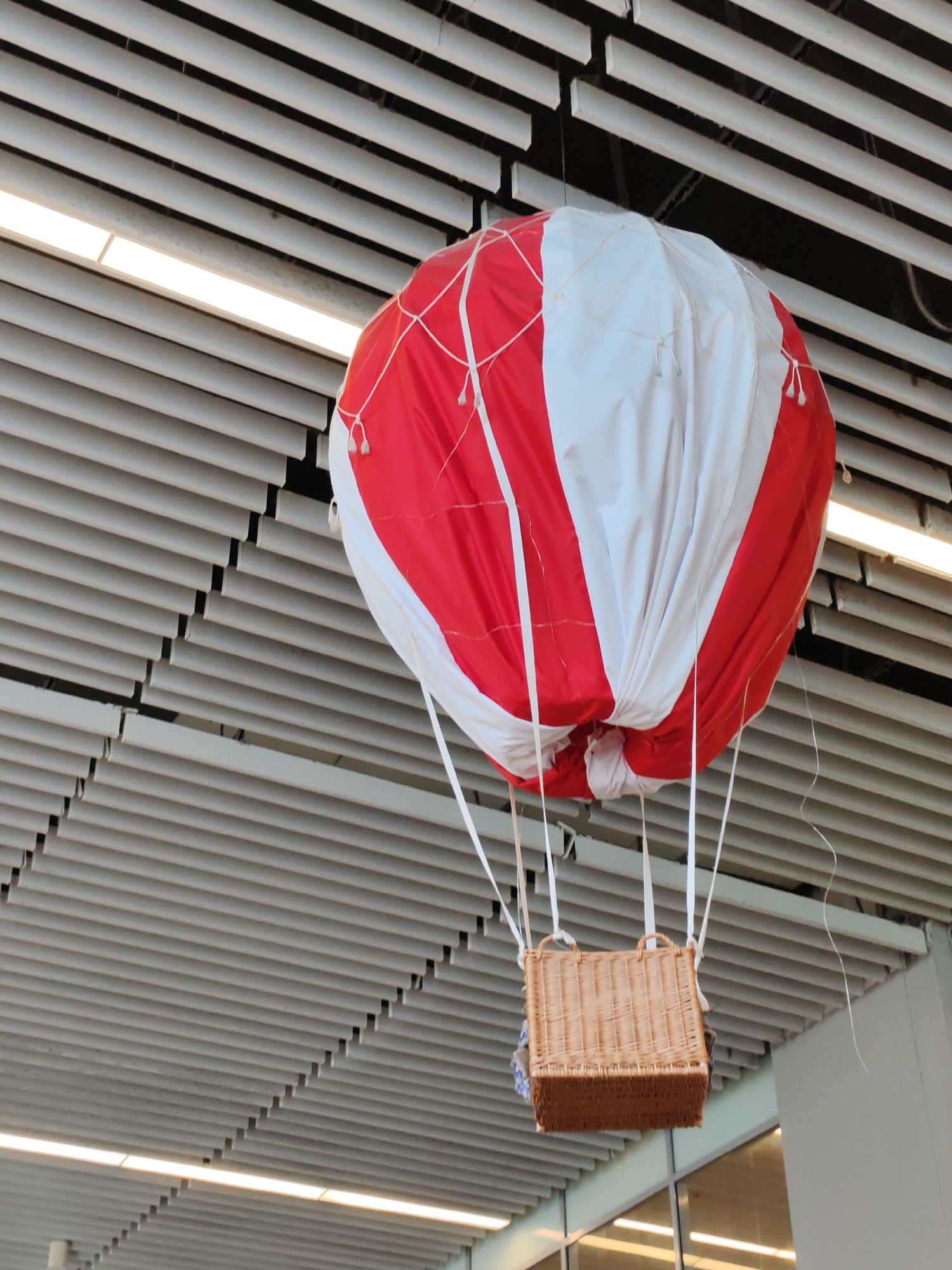 A decorative red and white hot air balloon with a woven basket hangs suspended from a slatted ceiling. The balloon is made of fabric panels and is secured by thin white ropes. The basket, made of wicker, hangs below the balloon. The ceiling is made of horizontal white slats with a fluorescent light fixture running horizontally across it. The image appears to be taken indoors, possibly in a public space. The location in Khrabrovo, Russia is not ascertainable from the image alone.