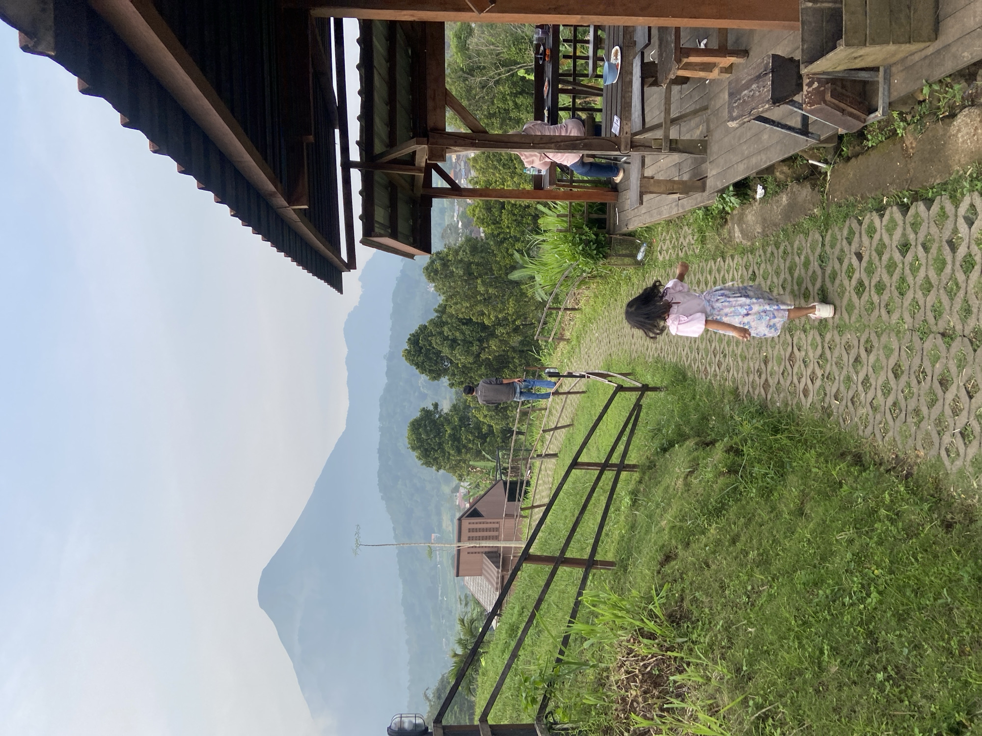 The scene is an outdoor area in Tamiajeng, Indonesia, featuring a mountainous backdrop under a hazy sky. A young girl in a light pink shirt and floral-patterned skirt walks on a gray-patterned concrete path. The path is bordered by green grass and leads towards a wooden gazebo or deck structure where a person is seated. A metal railing runs along the edge of a grassy area that slopes downward. Further down the slope, a building is visible amidst trees. Another person is walking on a pathway beyond the railing. The scene includes elements of nature (mountain, trees, grass) and built environment (gazebo, pathway, railing, building).