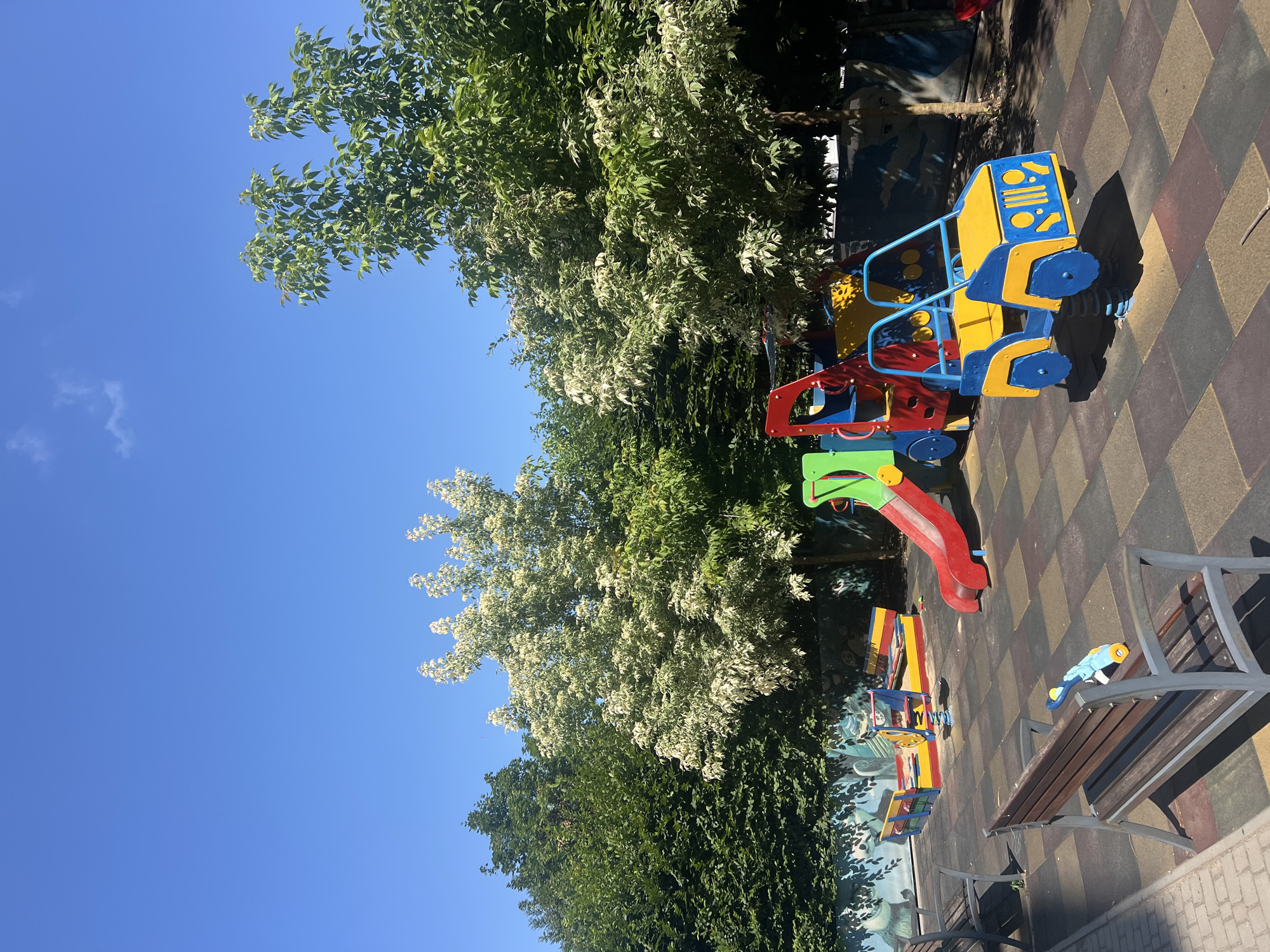 This is a wide, eye-level, outdoor shot of a colorful children's playground on a sunny day in Fontanka, Ukraine. The playground features a bright red slide, a blue and yellow spring-mounted car, and a sandpit with yellow and red accents. Several park benches are visible, one of which has a blue toy resting on it. The ground is covered with a patterned rubber matting in brown, beige, and dark gray. Lush green trees with some white blossoms frame the playground, and a clear blue sky with a single small cloud stretches above. A low, mural-painted wall runs along the back of the playground area. The image appears to be taken from a slightly elevated position, looking down onto the playground.
