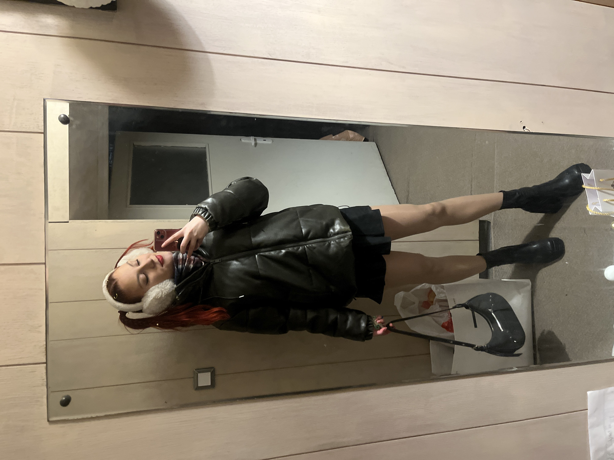 A woman is taking a selfie in a mirror. She is wearing white earmuffs, a dark puffer jacket, a short black skirt, and black boots. She is holding a black purse. The background shows a door, a shopping bag, and other indistinct items. The location cannot be determined to be Sofia, Bulgaria, from the given image.