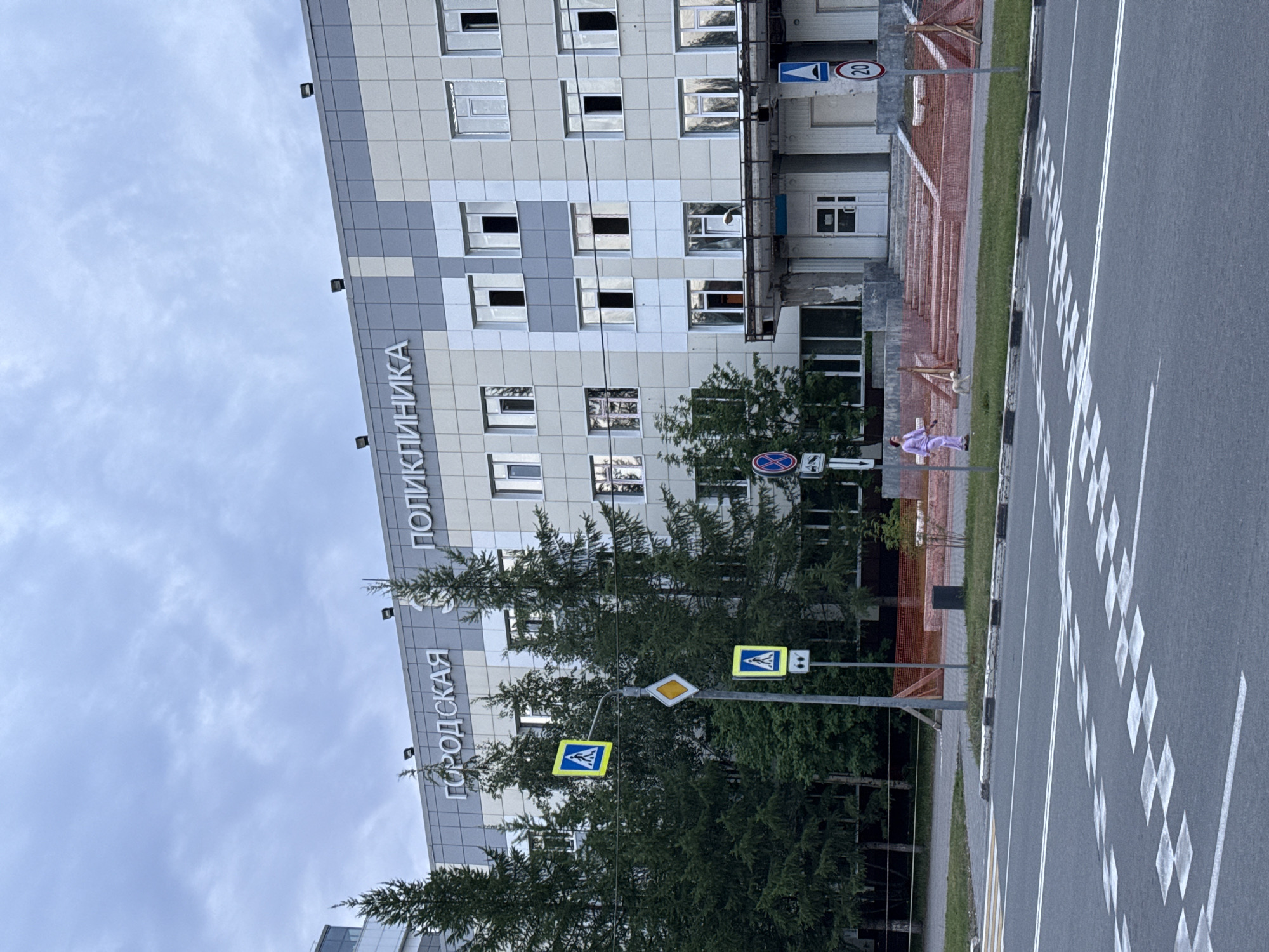 This image captures an outdoor scene, likely in Surgut, Russia, during the daytime under an overcast sky. The primary subject is a large, modern building with a facade of grey and white panels, featuring numerous windows. Prominently displayed on the building is Cyrillic text that reads "ПОЛИКЛИНИКА" (Polyclinic) and "ГОРОДСКАЯ" (City).
In front of the building, a tree-lined sidewalk runs parallel to a street. A person wearing a light purple outfit is visible on the sidewalk, walking and looking at their phone. A small dog accompanies them. The street itself has clear road markings, including a crosswalk with diamond patterns and white lines indicating lanes. Several traffic signs are present, including a pedestrian crossing sign with a blue background and a white pedestrian symbol, and a circular sign with a red border and the number "20" inside, likely indicating a speed limit. Another diamond-shaped yellow sign is also visible.
The scene suggests an urban environment with a medical facility as the central building. The weather appears to be cloudy, and the lighting is diffused, typical of an overcast day. The overall mood is neutral, depicting a normal day in an urban setting. The presence of the pedestrian signs and crosswalk indicates a public area with traffic and pedestrian activity.