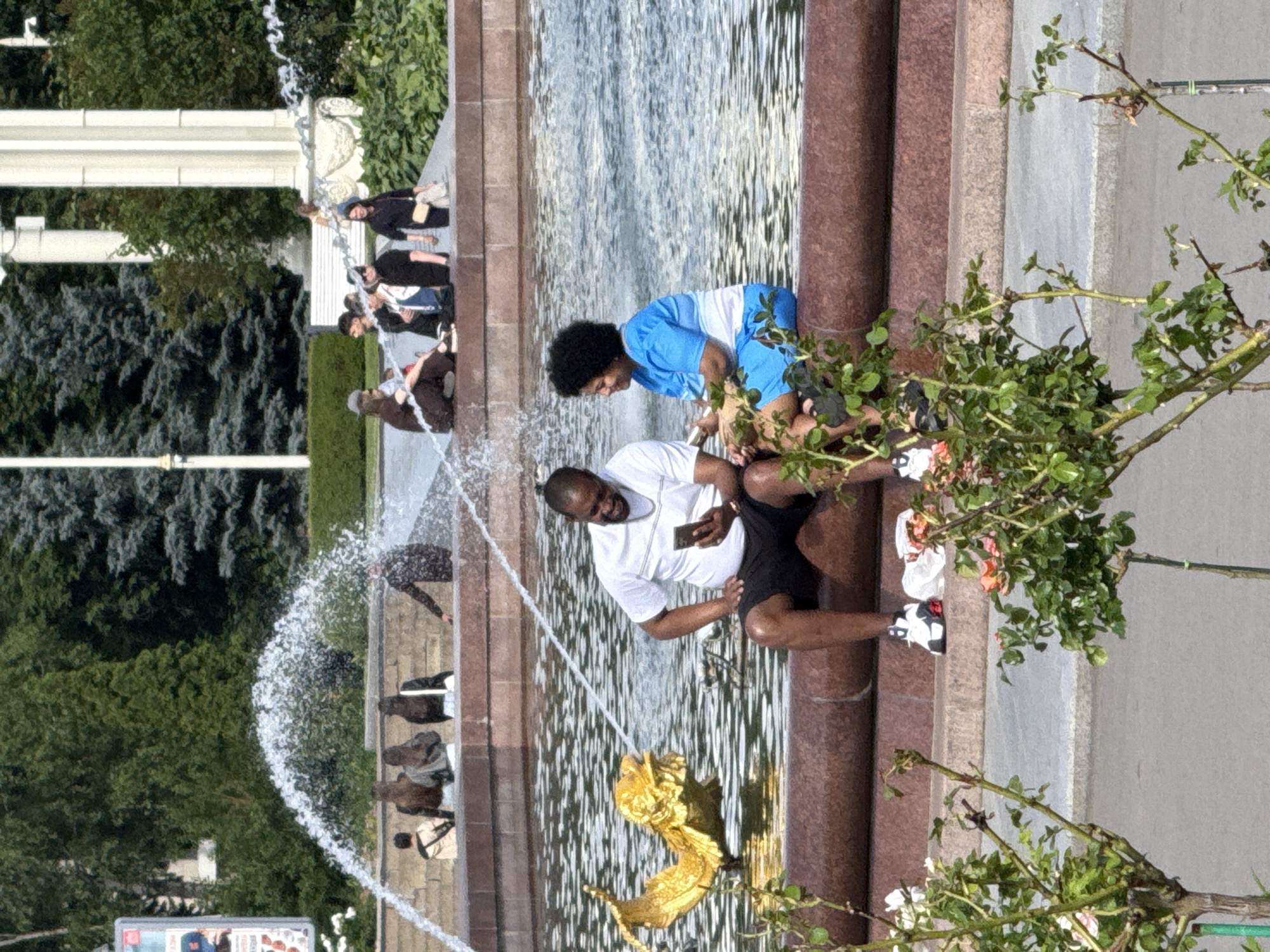 The image depicts a public fountain area, likely in Moscow, Russia, on a sunny day. The central subjects are several people interacting with the fountain. In the foreground, a man in a white t-shirt and black shorts is sitting by the fountain, holding a smartphone and appearing to be engaged with it. To his left, a woman in a blue and white outfit is leaning over the fountain, possibly interacting with the water or a decorative element. Other individuals are visible in the background, some sitting on the fountain's edge or standing nearby, suggesting a casual gathering or a place for public relaxation.
The fountain itself appears to be a multi-tiered structure with water cascading down. A prominent decorative element, a golden sculpted creature resembling a dragon or griffin, is visible within the fountain's basin. Lush green trees and foliage frame the scene, indicating a park-like setting. The time of day is suggested to be daytime by the bright lighting and the shadows cast. The mood appears relaxed and social, with people enjoying the outdoor space and the water feature. No specific text is clearly legible in the provided image, except for a partial view of a billboard in the lower left corner which is too blurry to decipher.
