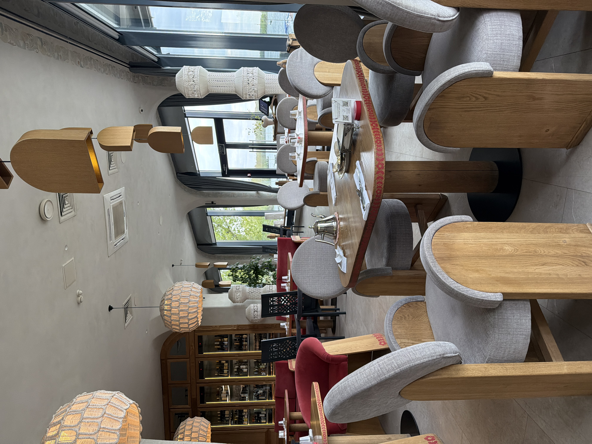 The image shows the interior of a restaurant in Krasnoyarsk, Russia. The restaurant has a modern design with curved tables, grey cushioned chairs with wooden arms, and decorative lighting fixtures hanging from the ceiling. Floor-to-ceiling windows offer a view of the outside. There's also a large wooden shelf, possible a wine rack. The dining area appears to be set up and ready for customers.