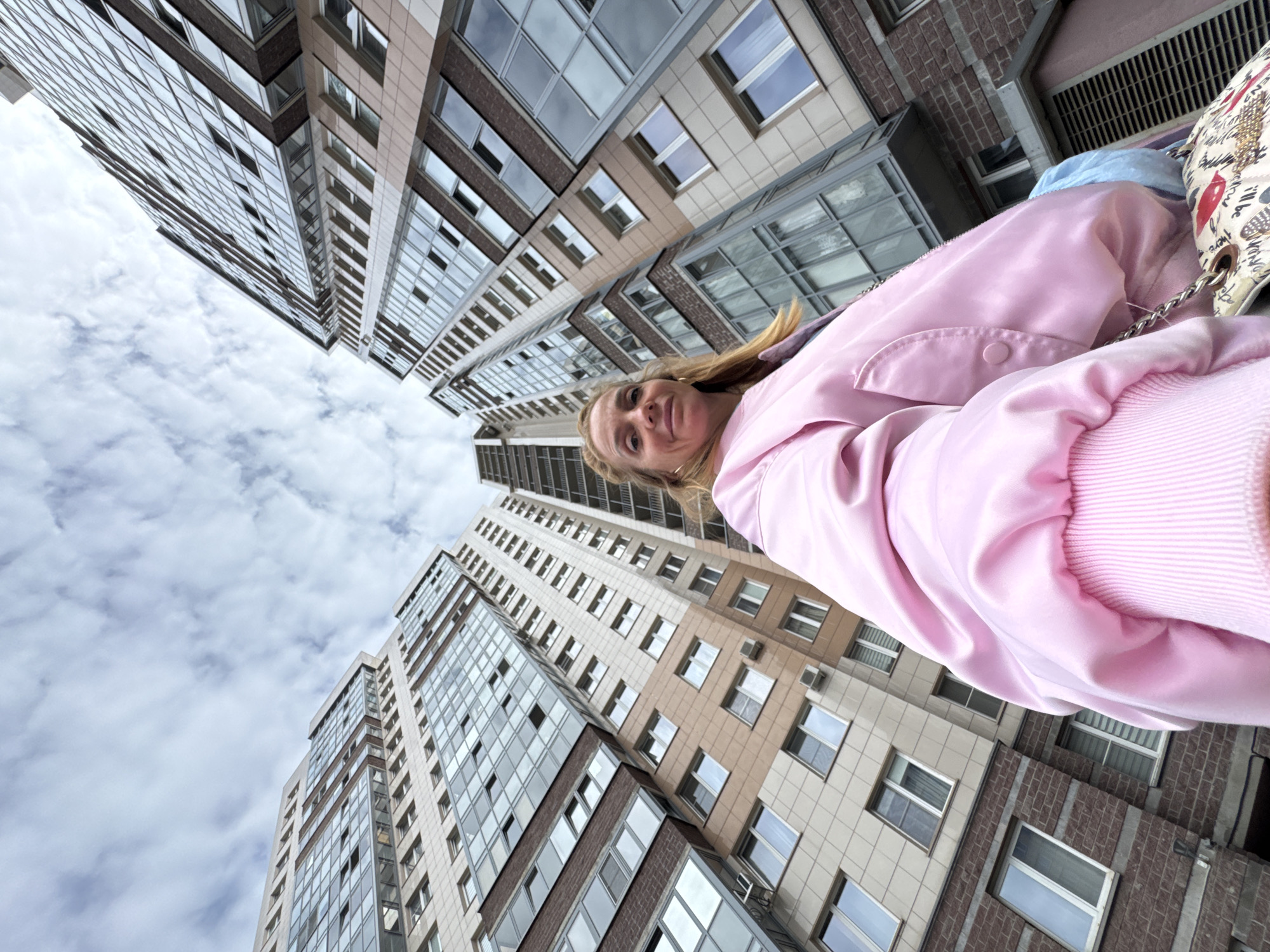 In Sinyavino, Russia, a woman in a pink bomber jacket stands in front of a modern apartment complex. The perspective is from a low angle, looking up at the towering buildings. The sky is cloudy with patches of blue. The woman is smiling and looking at the camera, holding a purse with a patterned design. The buildings are tall and contemporary, with many windows and balconies.