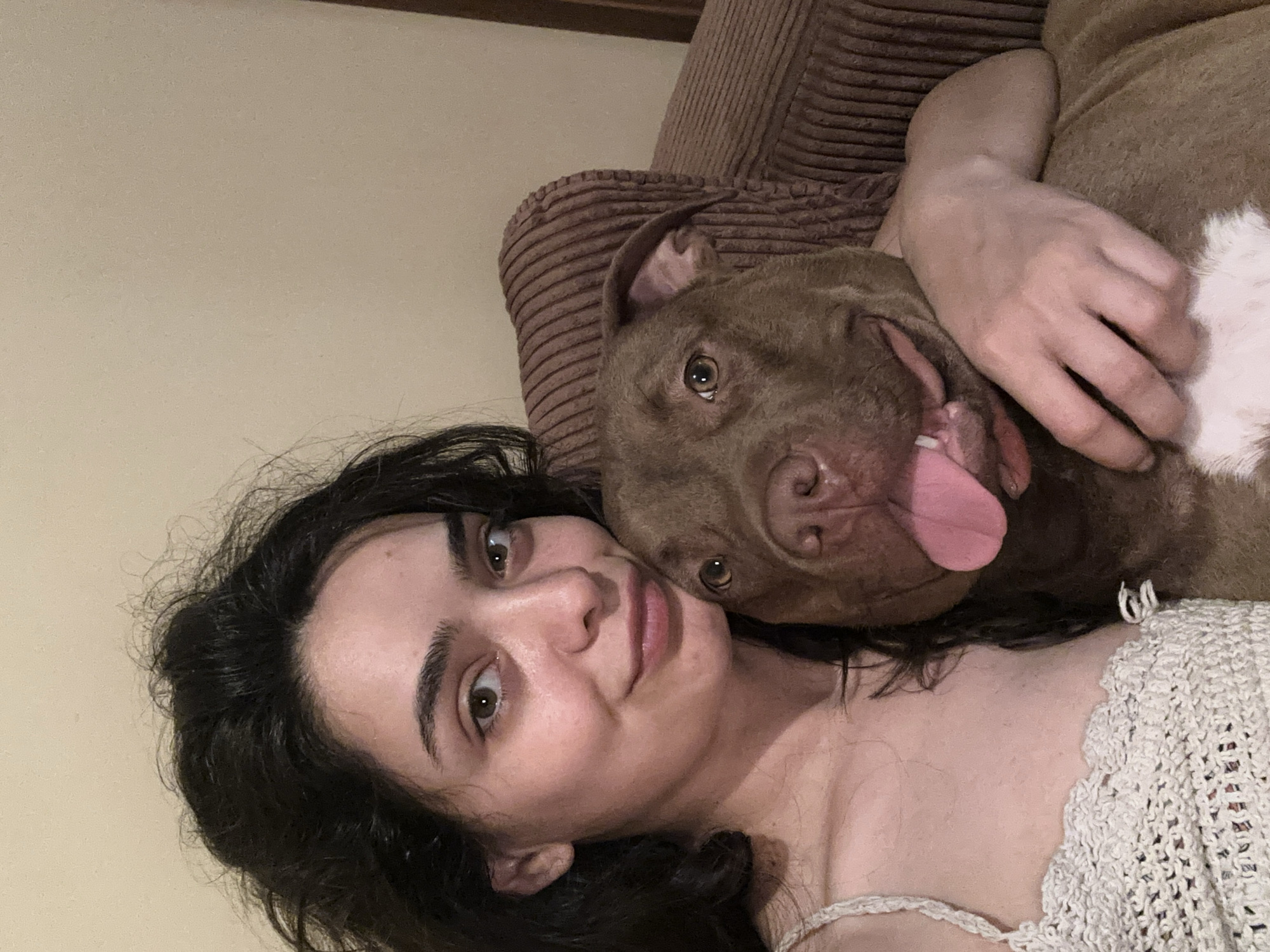 In the city of Malevo, Bulgaria, a young woman with dark, wavy hair and light skin is pictured with a brown pit bull dog. The woman is wearing a white, crocheted top and is looking towards the camera with a slight smile. The dog is resting its head on her shoulder and appears to be panting, with its tongue visible. They are both seated on a brown, corduroy-textured sofa. The background consists of a plain, light-colored wall. The lighting suggests an indoor setting, possibly a living room.