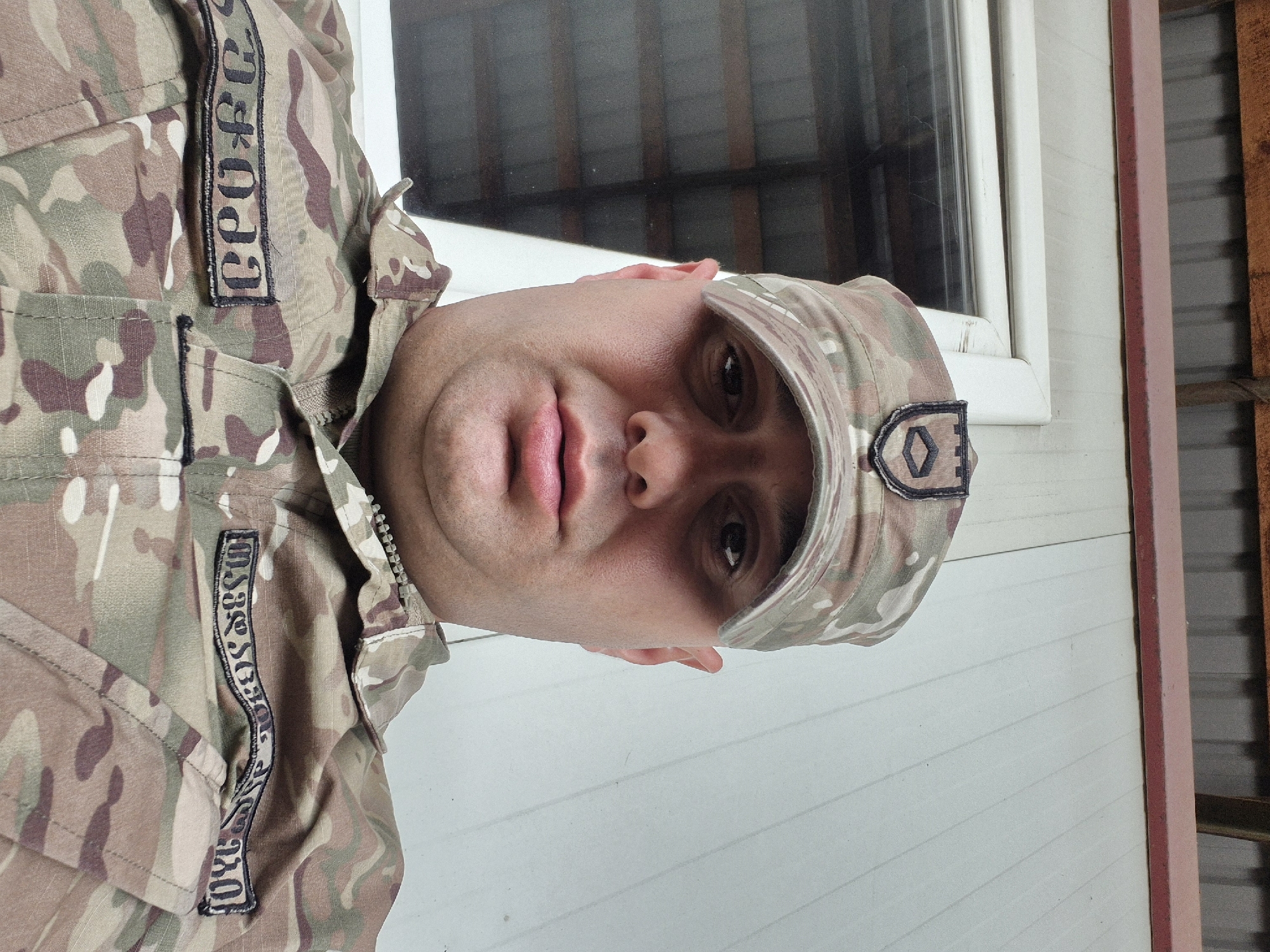 A close-up portrait captures a man in military attire. He wears a camouflage uniform and cap, both patterned in muted greens, browns, and tans. Patches on the uniform indicate military affiliation, one displaying the text "CRO&CF" and another "US Army". The man's face features a strong jawline, dark eyes, and full lips. He appears to be facing the camera directly. The background consists of a white-framed window and an exterior wall with horizontal siding painted white. A portion of the wall's trim is painted pink. The location is likely Tbilisi, Georgia, given the Cyrillic character in the military patches. The setting appears to be indoors, potentially near the exterior of a building.