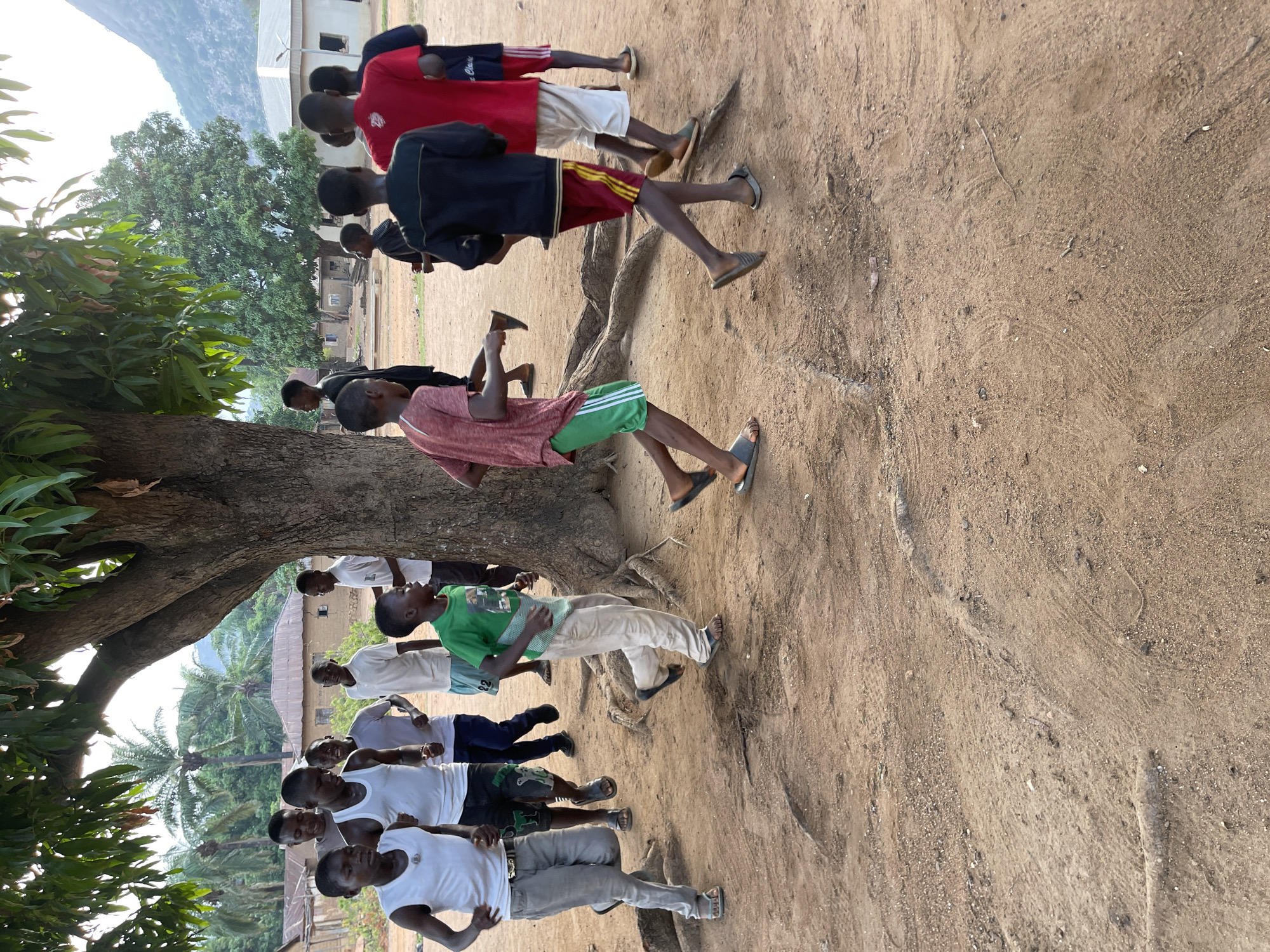 A medium-angle shot captures a group of at least thirteen people, presumably young men or adolescent boys, in Tabu, Nigeria. The scene is outdoors, on a dirt ground, with a large tree in the midground. The people are arranged in a loose, slightly staggered line. They are wearing varied clothing, including t-shirts (white, green, and red), shorts (red, green, and white), pants (tan and gray), and footwear like sandals. The tree's trunk is substantial, with exposed roots visible above the ground. In the background, buildings and additional trees are visible, including a palm tree. A hill or small mountain can be seen in the distance. The overall lighting suggests daytime.