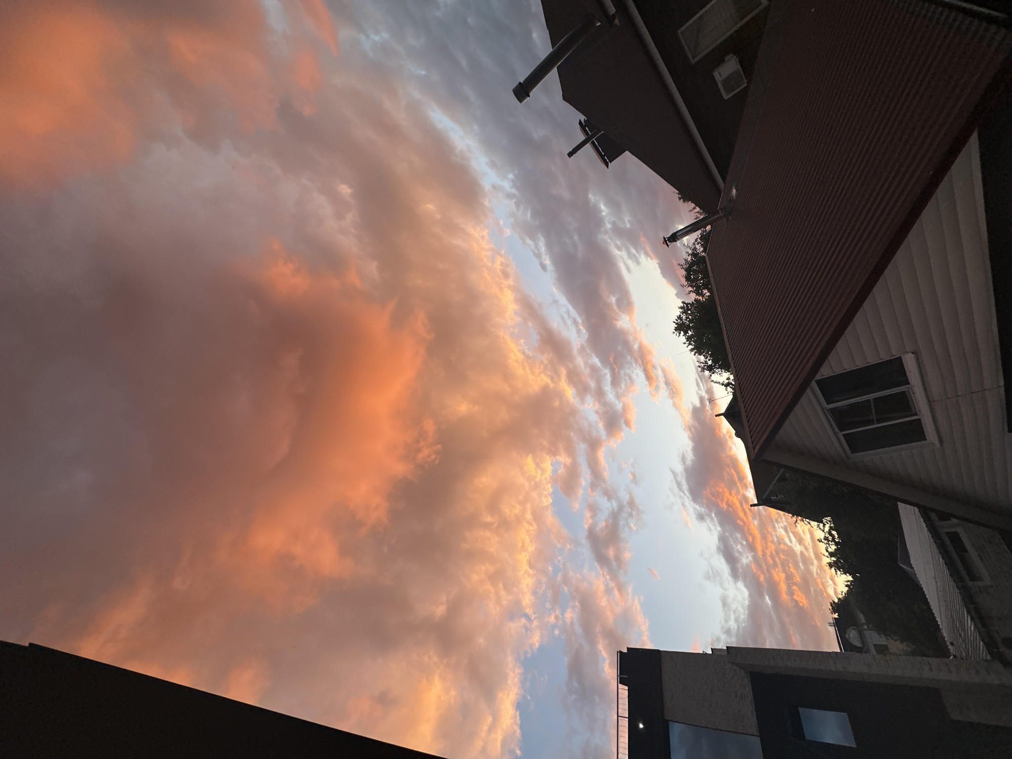 The image depicts a cloudy sky at sunset, with hues of orange and pink dominating the clouds. In the foreground, there are several rooftops of houses and a modern building on the left. The architecture suggests an urban or suburban setting. The specific location is the city of Nizhny Novgorod, Russia, as indicated by the provided metadata. There are no people visible in the image, nor are there any discernible actions or interactions. The scene is static and captures a moment in time with the focus on the interplay of natural light and man-made structures.