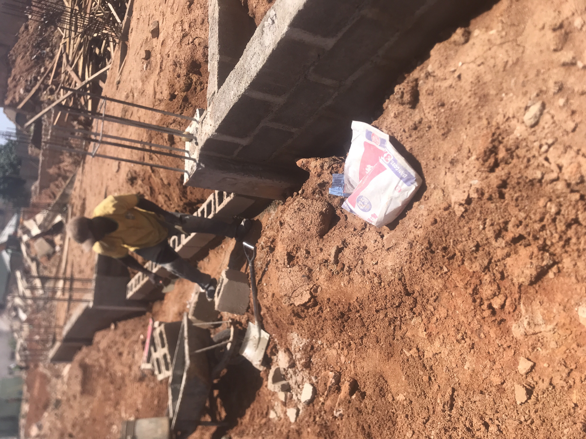 A construction site in Jos, Nigeria is shown. A worker in a yellow shirt is interacting with concrete blocks and metal rebar. A bag of Dangote cement is sitting on the dirt ground. The scene includes a shovel and other construction-related materials. The ground is red earth. The image appears to be taken at an angle, possibly by a hand-held device.
