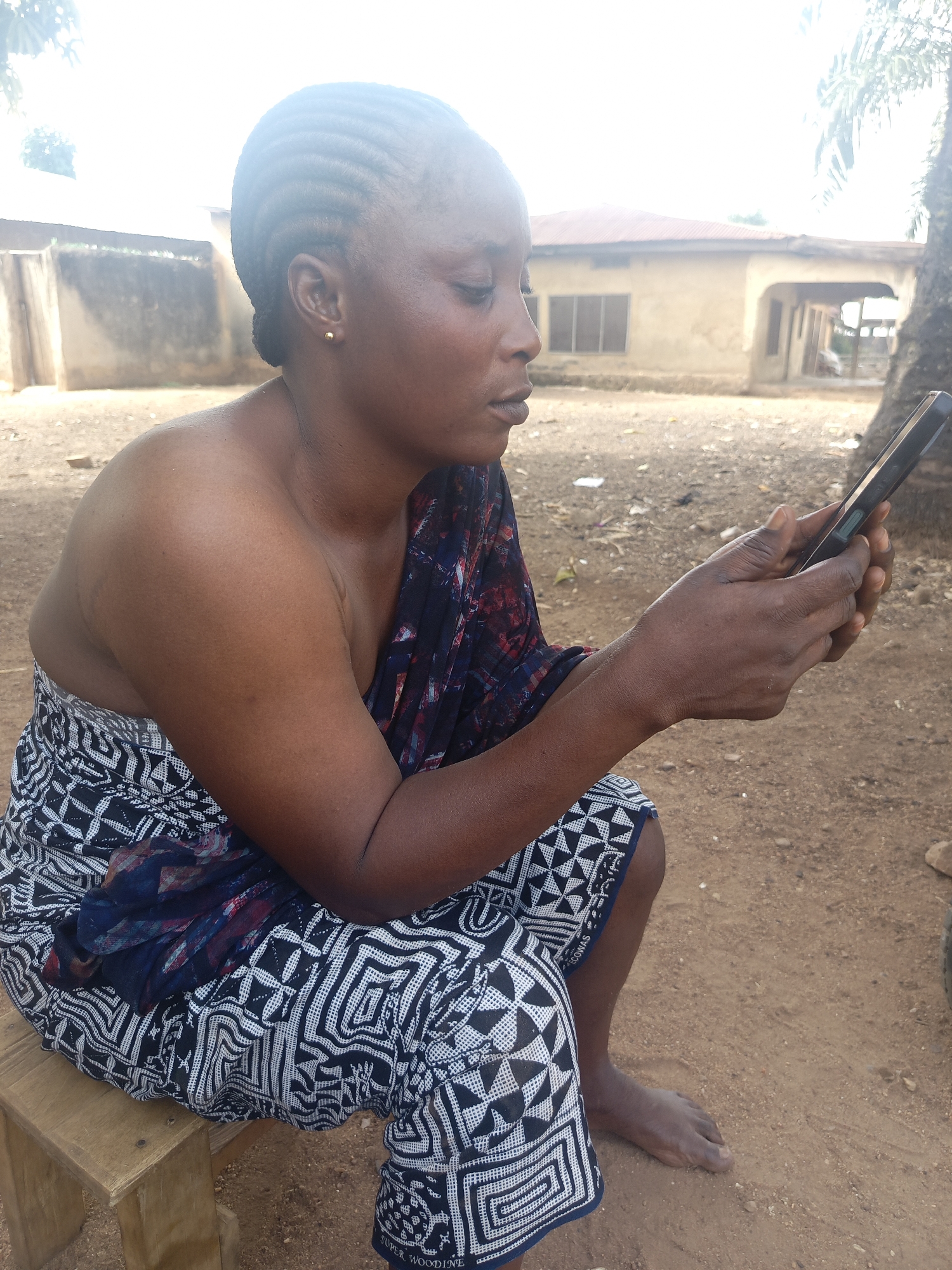 An adult woman with dark skin and braided hair, wearing a patterned garment and a shawl, sits on a small wooden stool. She is holding a smartphone and looking at it intently. The setting appears to be outdoors in Akwanga, Nigeria, with a dirt ground and a building in the background. A tree is also visible to the right of the frame.