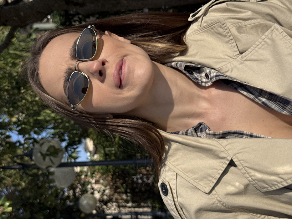 An outdoor, sunlit, mid-shot portrait features a single adult female in Sirmione, Italy, likely in a park or promenade area. The woman, positioned slightly off-center, is illuminated by direct sunlight from the upper left, casting shadows. She has long, dark brown hair and wears narrow, silver-rimmed, oval-shaped sunglasses with dark lenses. Her eyebrows are slightly furrowed, and her lower lip is partially tucked under, creating a specific expression. She is dressed in an unbuttoned light beige trench coat over an open-collared plaid shirt with blue, white, and brown patterning, revealing a portion of her chest. In the blurred background to the left, two dark, period-style streetlights with spherical white lamps are visible. Behind these, out-of-focus green foliage from trees and a faint, light-colored building are discernible against a clear blue sky.