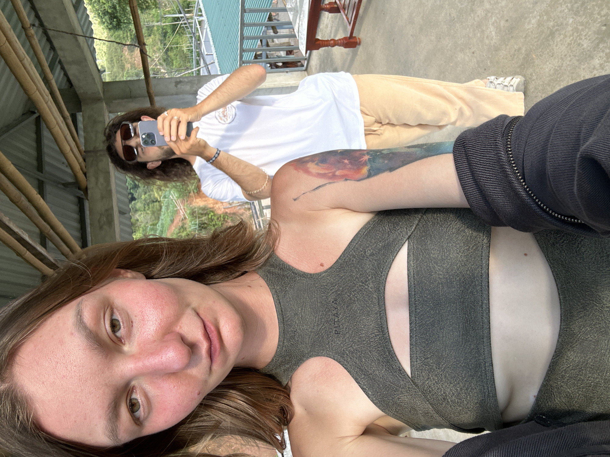 The image depicts two individuals on an elevated outdoor viewing platform or balcony in Tú Lệ, Vietnam, characterized by a visible roof structure overhead and a mountainous, green landscape in the background.
In the foreground, a woman with medium-length brown hair is taking a selfie. She has light skin with some redness on her cheeks and nose, and her lips are slightly parted in a neutral expression. She wears a dark olive green athletic top with a cutout revealing her upper chest and midriff, and the text "PITAYA" is visible on the chest area. Her left arm features a prominent, colorful tattoo spanning the bicep and shoulder, depicting an abstract, fiery landscape. Part of a dark, zippered sleeve is visible on her lower right.
In the mid-ground, slightly behind and to the right of the woman, a man with long, dark, wavy hair stands. He wears dark rectangular sunglasses, a white short-sleeved t-shirt with a graphic logo on the chest, and light beige or khaki loose-fitting pants. He has multiple beaded bracelets on his right wrist and a silver-colored ring on his left ring finger. He is holding a grey smartphone with both hands, aiming it away from the viewer, appearing to capture the scenery. His footwear consists of light-colored, multi-strap sandals or shoes.
The setting includes a grey concrete floor and an overhead structure composed of concrete beams and lighter bamboo poles, supporting a corrugated metal roof. Beyond the railing on the right, a small, reddish-brown wooden table with a lighter top is visible. The background reveals a lush, mountainous terrain with dense green vegetation and what appear to be cultivated fields or terraces on the slopes, consistent with the agricultural landscape of Tú Lệ. Another building with a green roof is partially visible in the distance. The scene is illuminated by natural daylight.