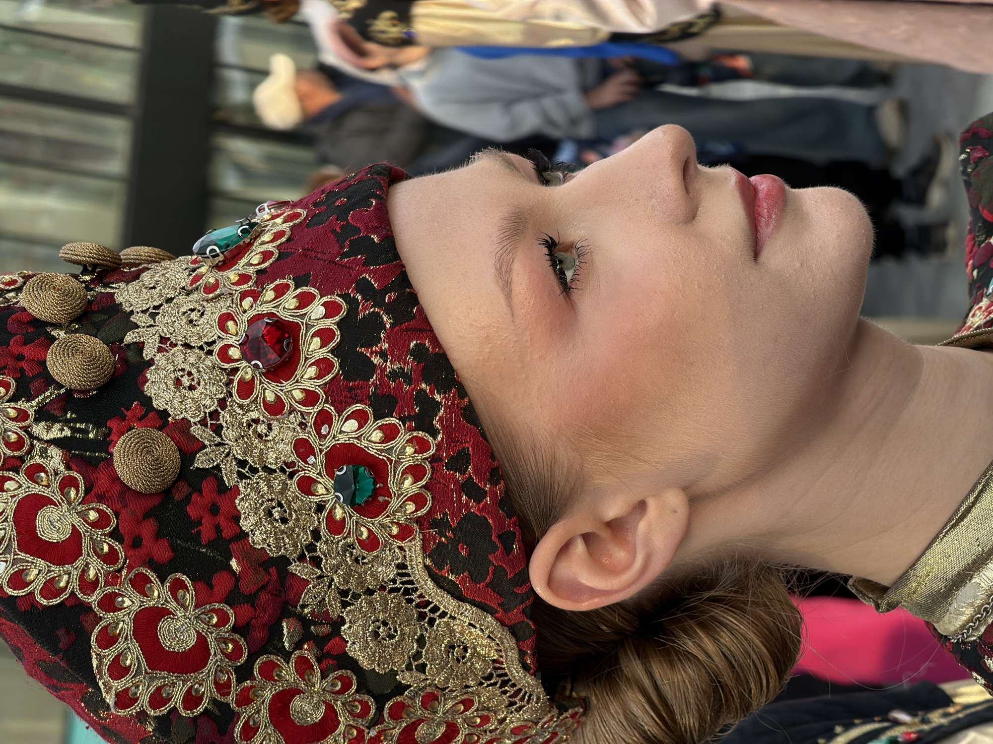 A young woman is shown from the side, wearing a highly ornate, traditional headdress with red, black, and gold embroidery, as well as red and green gemstones. Her hair is pulled back into a bun. She is wearing a matching ornate collar. The background is blurred but appears to be an outdoor public space, possibly a street or market, with other people visible in the distance, some wearing casual clothing and one with a baseball cap. The lighting suggests it is daytime.