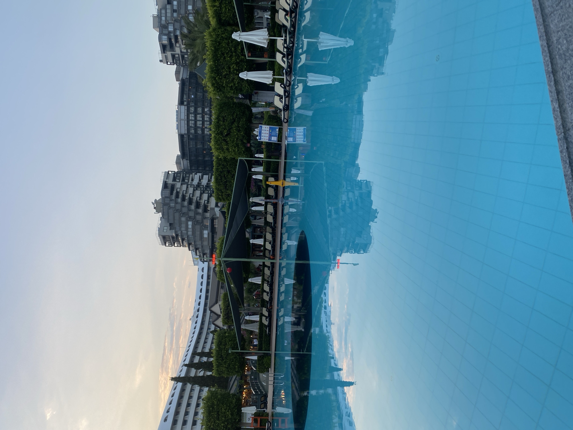 The image shows a resort swimming pool area in Aksu, Turkey, during what appears to be early morning or late evening. The sky is clear with a gradient of light blue and a hint of pinkish clouds. A large, empty swimming pool with blue tiled bottom occupies the foreground, its surface reflecting the sky and surrounding structures.
To the left and in the background, a large, curved white hotel building is visible, with multiple levels and arched windows. To the right of this building, and further into the background, are modern, multi-story hotels with grey facades and numerous windows. Some are angular, while one in the center has a more circular design. Green trees and hedges are strategically placed around the resort.
In the middle ground, partially obscured by the pool's reflection, are rows of empty lounge chairs and white umbrellas set up under dark green canopies. A few tall, slender trees are visible behind the hotel on the left. One structure with a red digital display showing "13:50:03" is visible near a dark green canopy. A yellow cone and some signage are also present. The overall atmosphere is tranquil, with no people visible in the frame, suggesting a quiet moment before or after the resort's peak activity.