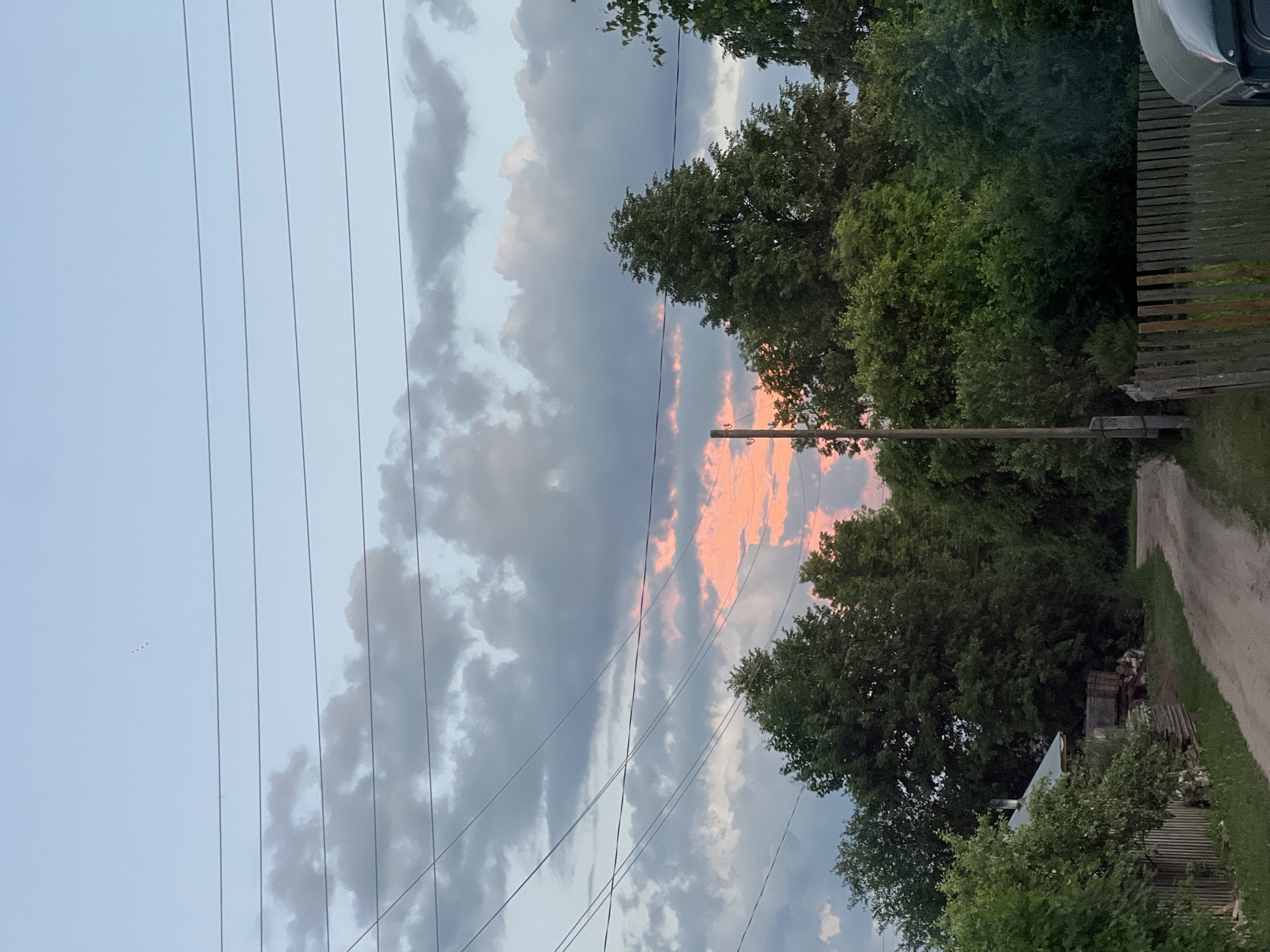 The image shows a rural road in the village of Pigozino, Russia, leading towards a dense line of trees. A power line with multiple wires stretches across the upper portion of the frame. A small, rustic house with a metal roof and a chimney is visible on the left side of the road, partially obscured by foliage. A wooden fence runs along the right side of the road, and a utility pole stands near the left edge. In the sky, a band of soft pink and orange clouds is visible through gaps in darker grey clouds, indicating either sunrise or sunset. A few small, pinkish dots, possibly distant kites or balloons, are visible in the upper left quadrant of the sky. The overall scene depicts a quiet, natural environment.