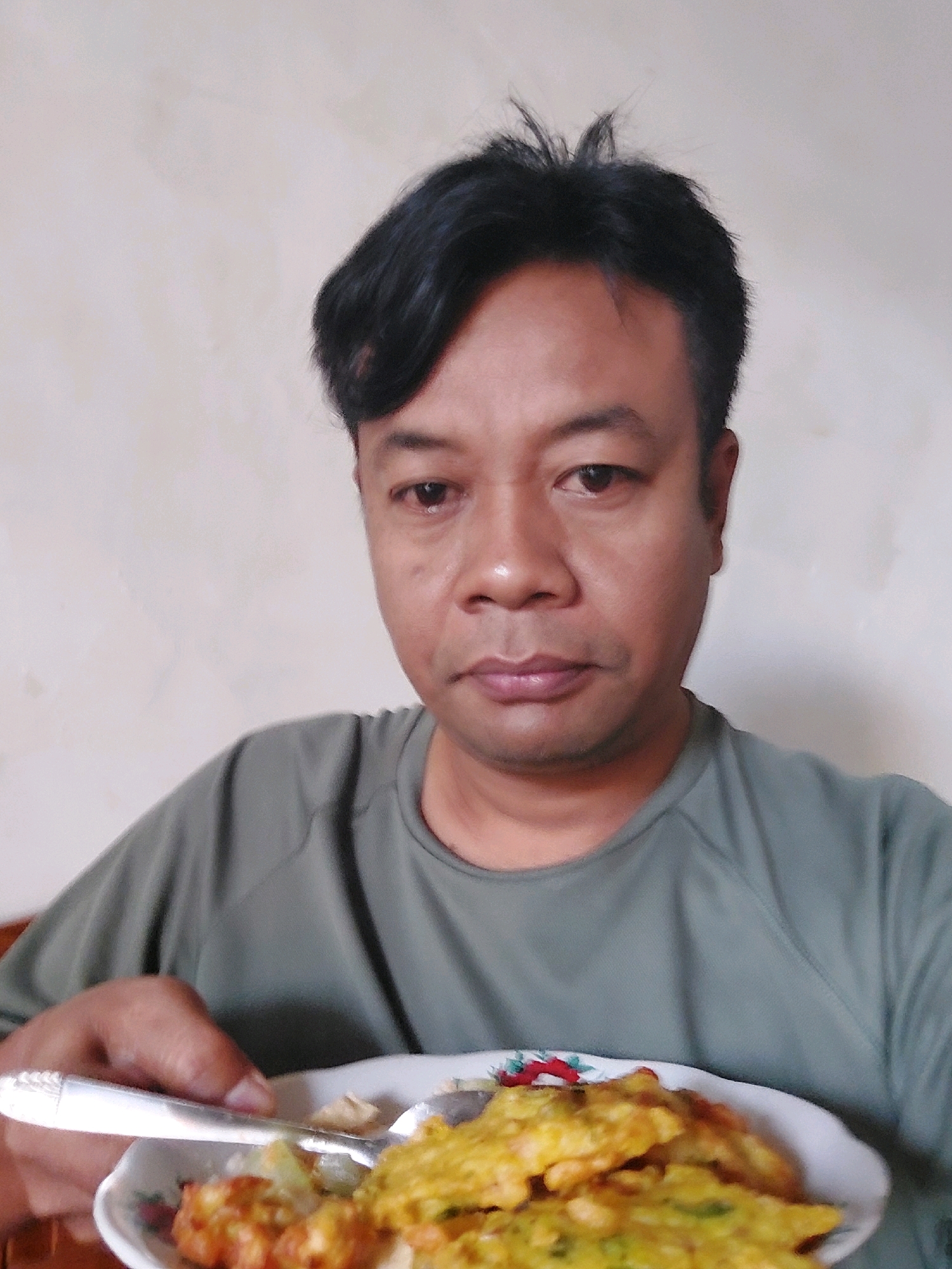 A medium shot, taken from a slightly high angle, captures an adult male with dark, tousled hair and medium-toned skin. He is wearing a dark green t-shirt and is looking directly at the camera with a neutral expression. The background is a plain, lightly textured cream-colored wall. In the foreground, he holds a white plate containing food with a silver fork and spoon. The food appears to be fried, with visible chunks of what looks like vegetables or fritters. The image suggests he is in a casual indoor setting, possibly in Sarangan, Indonesia, where he is about to eat.