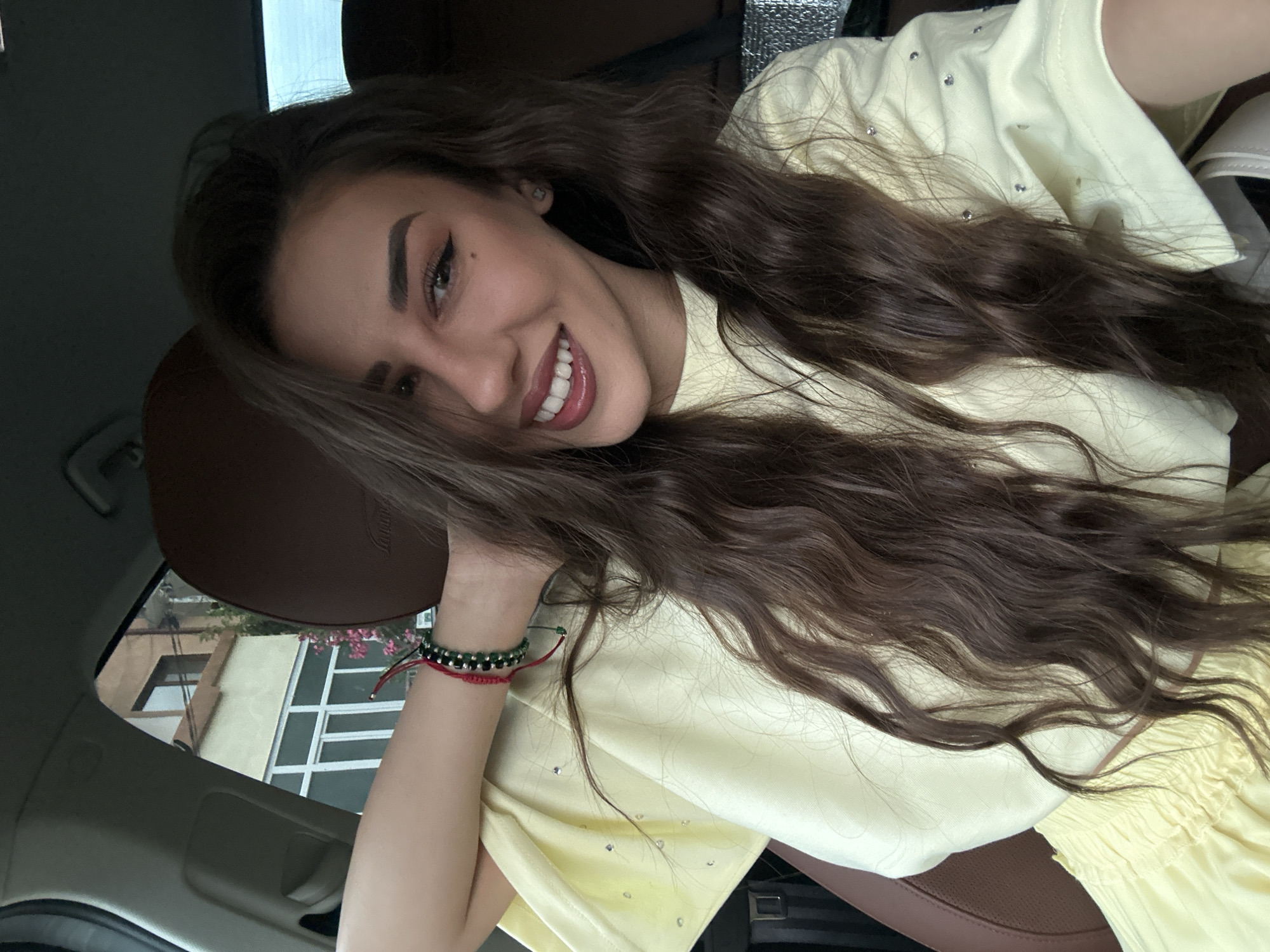 In the city of Petrich, Bulgaria, a young woman with long, wavy brown hair is pictured inside a car. She is wearing a light yellow top adorned with small, shimmering embellishments. Her left arm is resting behind her head, revealing a bracelet on her wrist made of green and red beads. The woman is looking towards the camera with a wide smile, her lips colored in a glossy, deep rose shade. Her eyes, accented with winged eyeliner, convey a friendly expression. The interior of the car features brown leather seats, and the background shows a building visible through the car window, suggesting an urban or suburban setting.