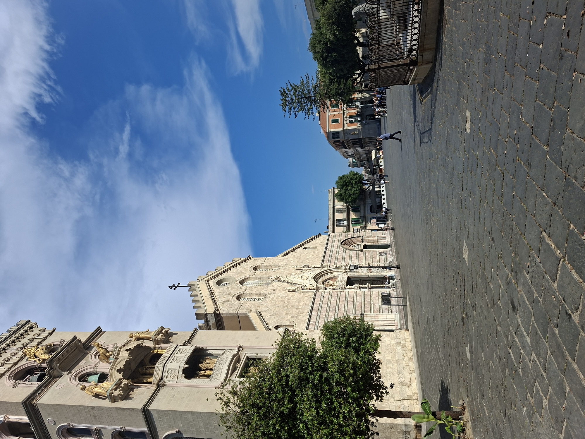 The image shows the Messina Cathedral in Messina, Italy. The cathedral is constructed of stone and has a facade with sculptures and religious iconography. A tall bell tower with golden statues is visible on the left side. A cobblestone square stretches out in front of the cathedral. A few people are seen walking in the distance. The sky is blue with scattered clouds. Trees are present in the image, positioned between the cathedral and the foreground.
