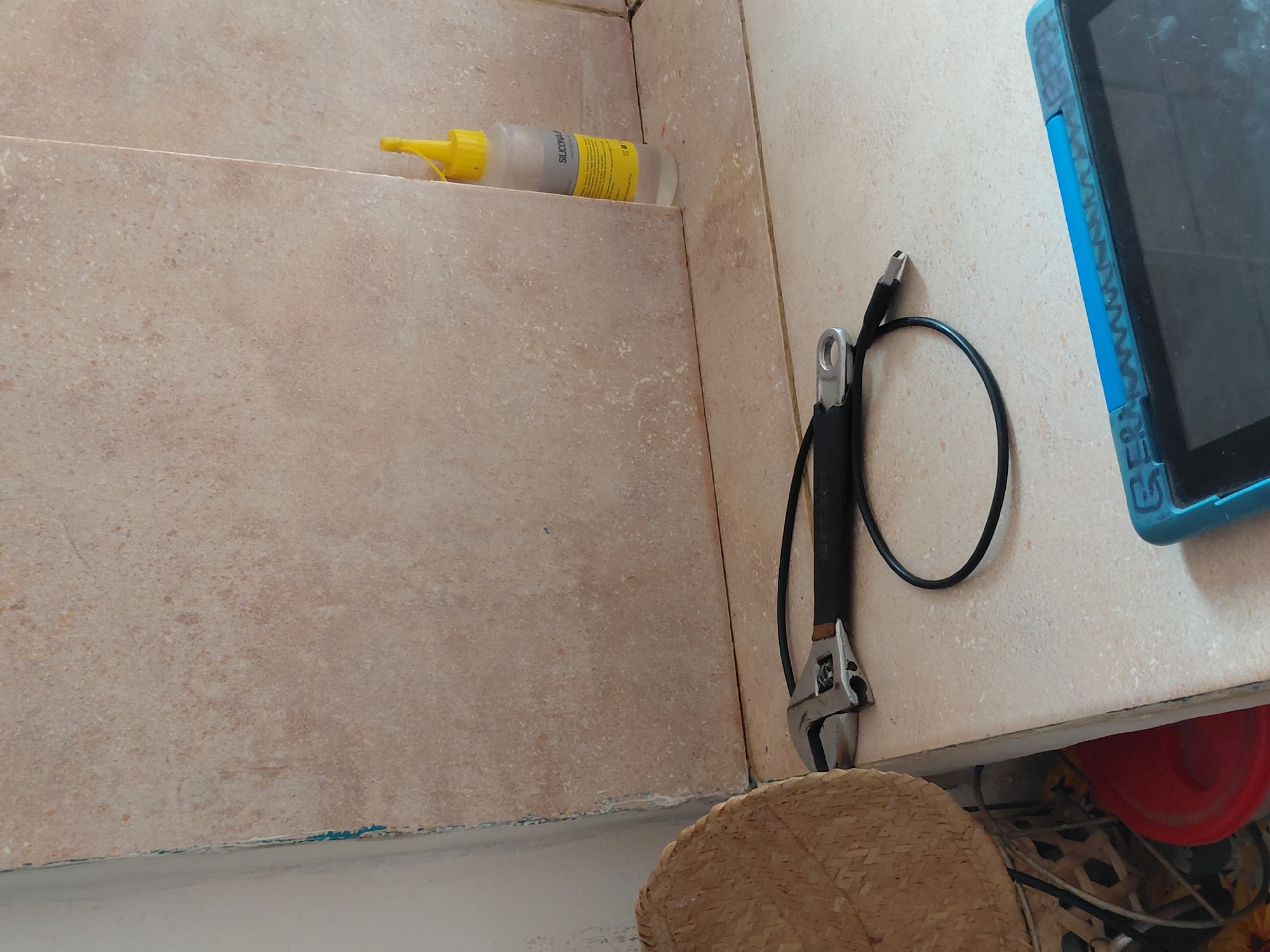 The image displays an indoor corner, likely a bathroom or kitchen, characterized by light beige, mottled-patterned tiles on both vertical and horizontal surfaces.
On an upper tiled ledge to the left, a clear plastic bottle with a bright yellow pointed cap is positioned. The bottle contains a white substance and its label partially shows the text "SILICON-J," indicating it is a type of sealant or adhesive.
On a lower tiled counter to the right, an adjustable wrench with a black handle is seen. It is hooked to a coiled black cable or hose. Adjacent to the wrench and cable, a tablet computer in a vibrant blue protective case is partially visible, showing its textured side and a segment of its dark screen.
In the bottom right corner, a portion of a woven straw item, resembling a hat or basket, is present. Below the counter, a curved red plastic object, possibly a bowl or bucket, can be faintly seen.
No specific activity is taking place, but the presence of tools like the wrench and sealant suggests that maintenance, repair, or a DIY project might have recently occurred or is pending in this space. The lighting indicates it is daytime. The scene portrays a functional area within a domestic setting.